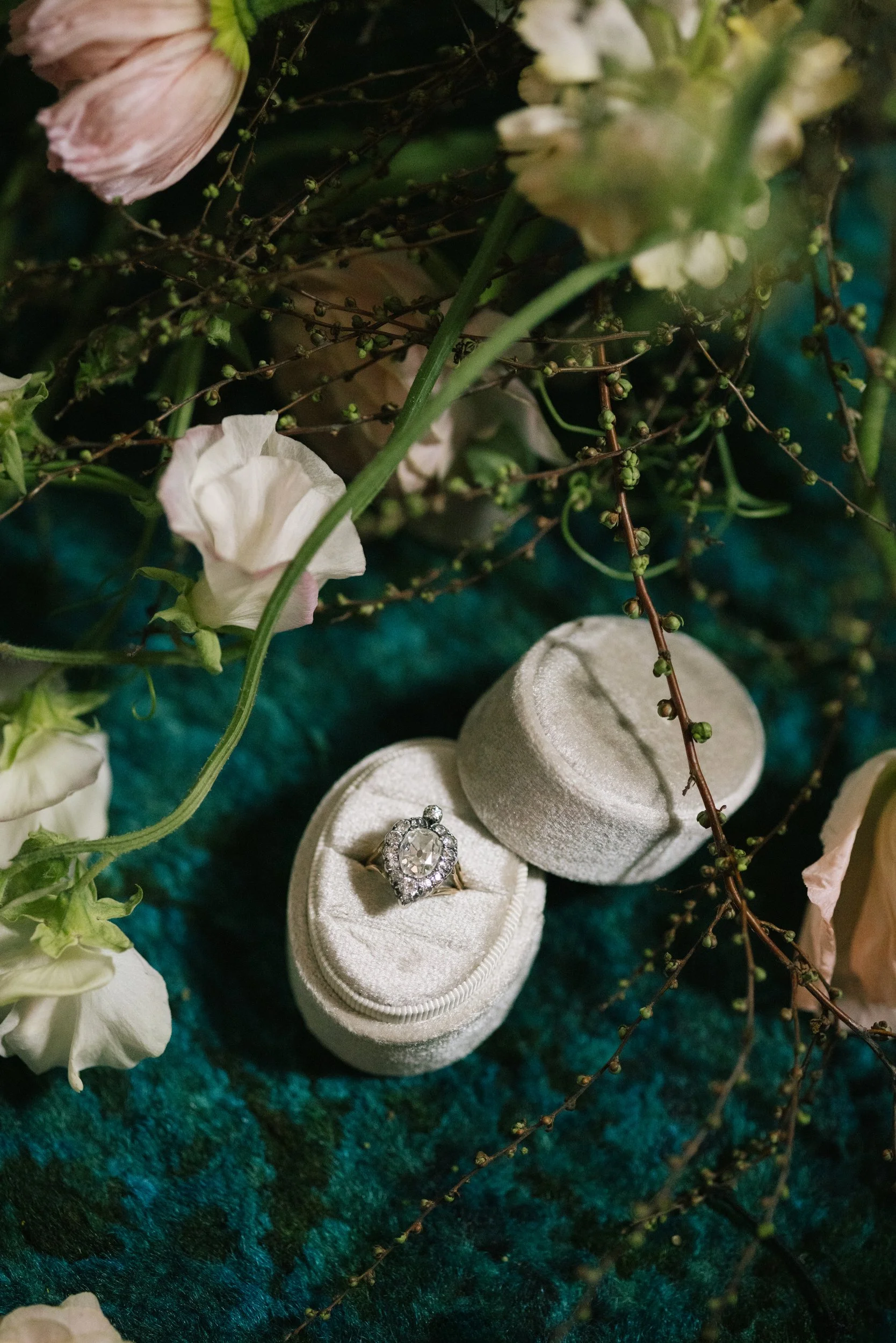Close-up of a jewelry ring in a white velvet ring box, surrounded by white and pink flowers and some sprigs with small green berries, on a dark green textured surface.