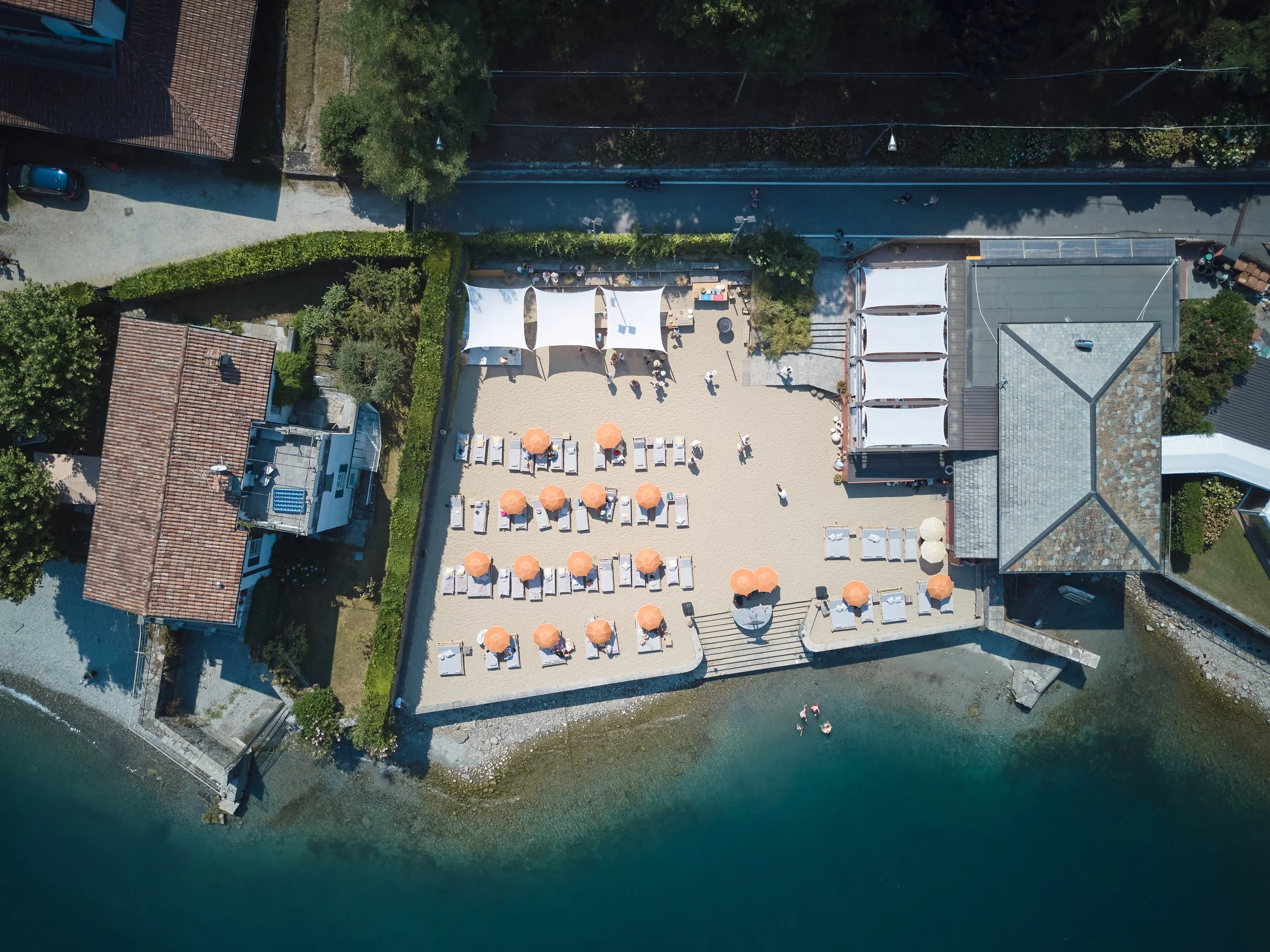 An aerial view of a waterfront property with a sandy area featuring white canopies, orange umbrellas, and lounge chairs, bordered by greenery and adjacent to a river with a dock and boats.