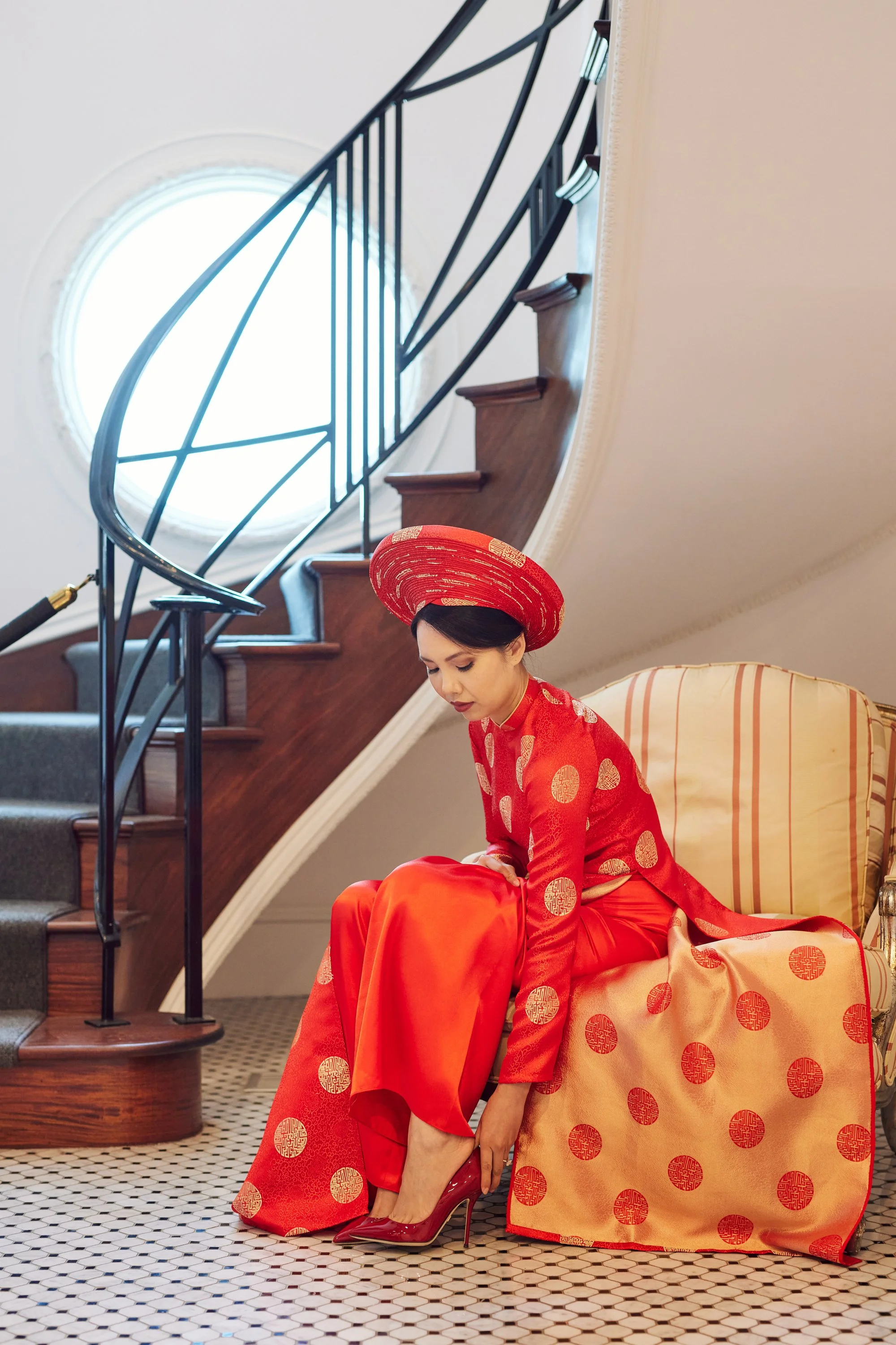 A woman dressed in a red and gold traditional Asian outfit, sitting on a yellow and orange chair, adjusting her red high-heeled shoe. She is under a staircase with black rails and a round window in the background.