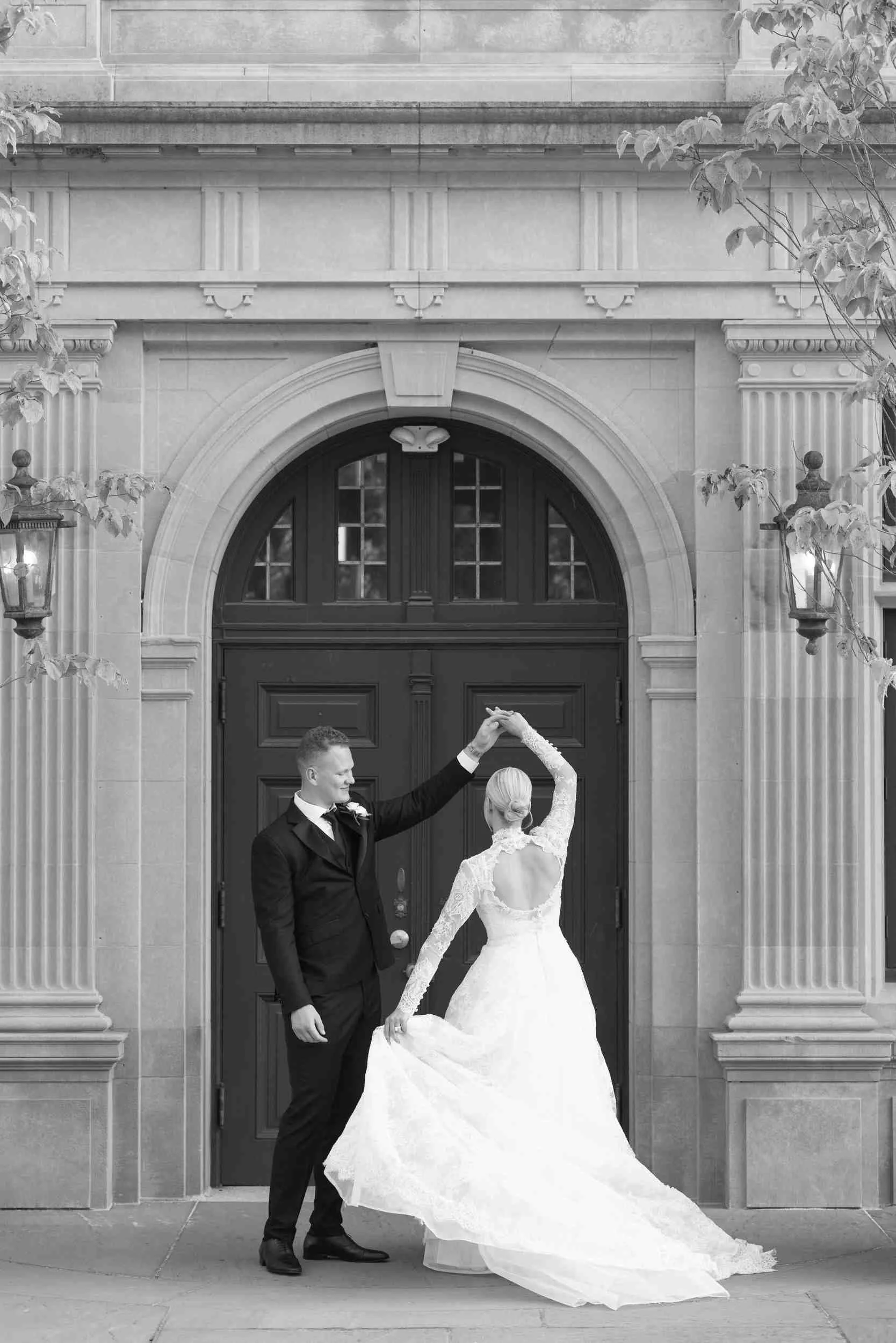 A bride and groom dance outside in front of a large wooden door and stone building, with the bride in a long white wedding gown and the groom in a black tuxedo, holding hands overhead.