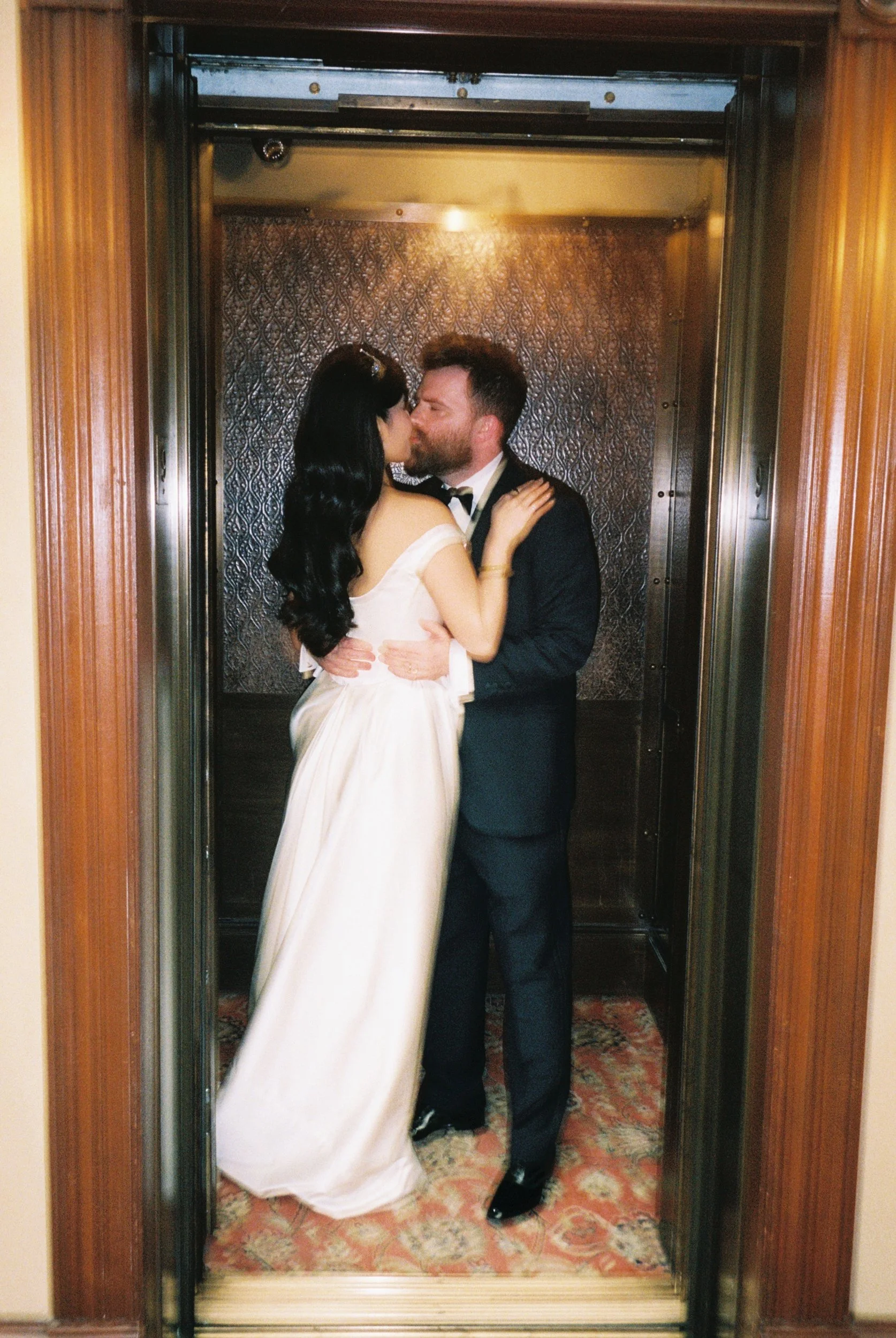 A man and woman kissing in an elevator. The woman wears a white dress, and the man wears a dark suit.