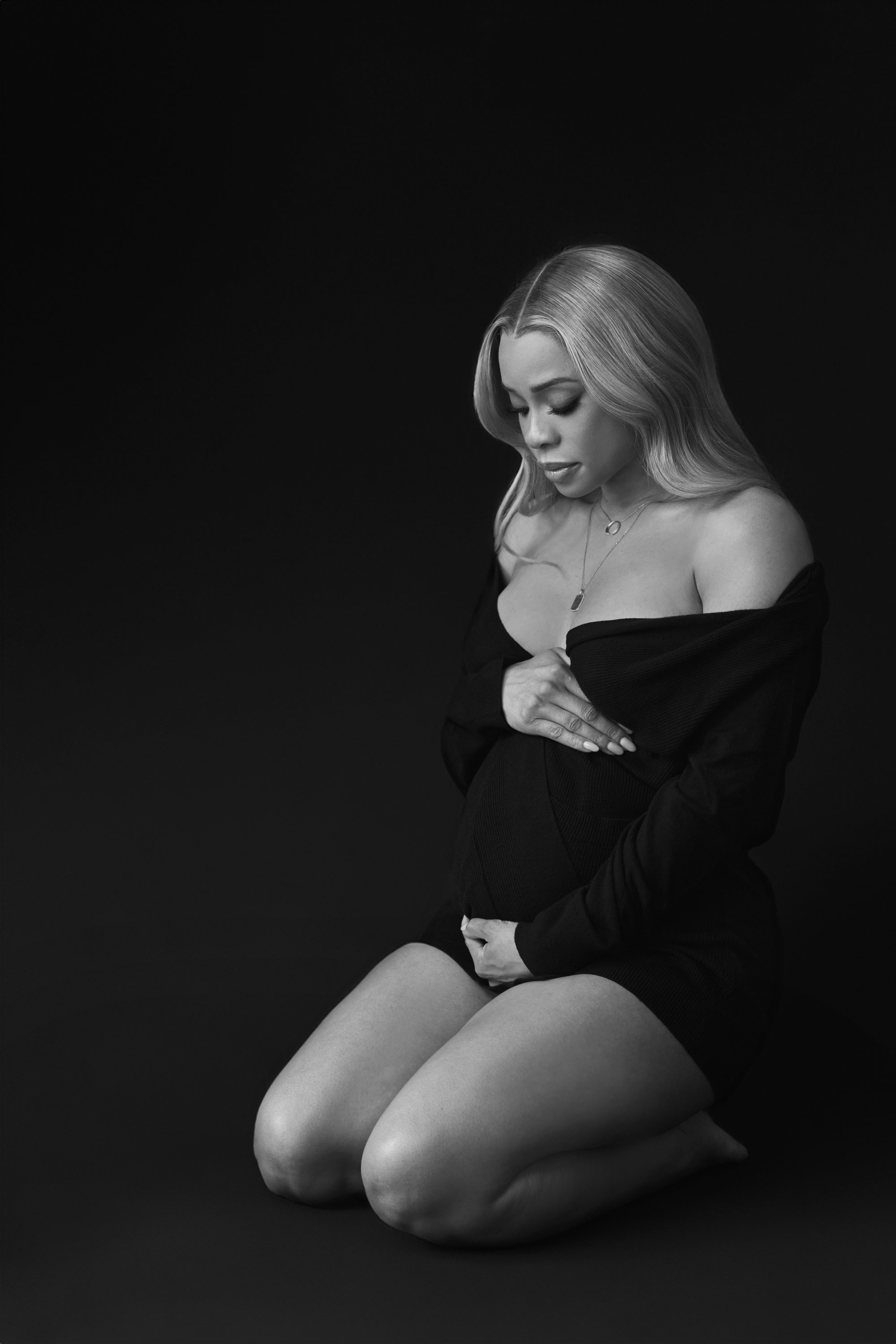Black and white photo of a woman kneeling with her head bowed and eyes closed, wearing an off-the-shoulder black top and jewelry, against a dark background.