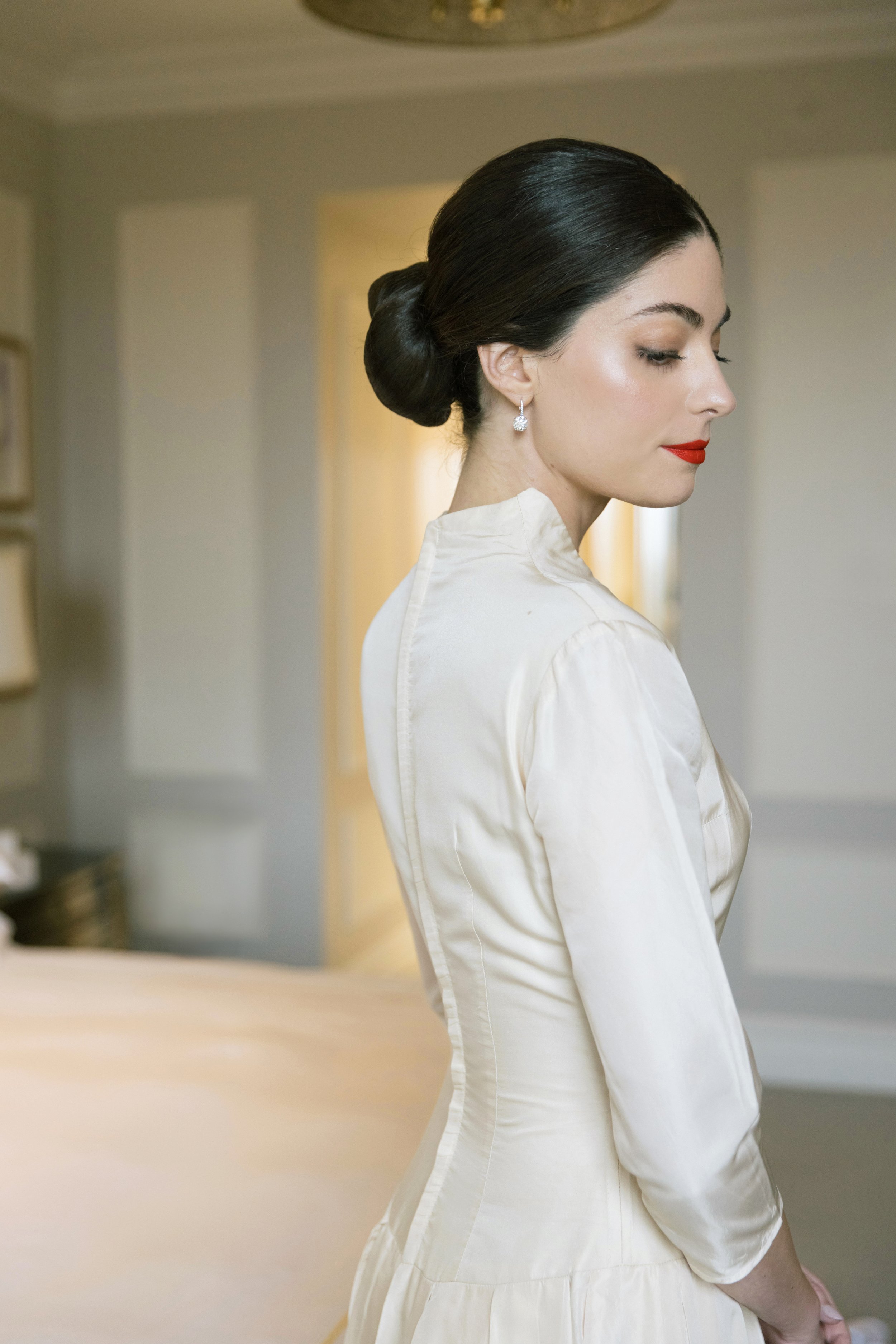 A woman with dark hair styled in a bun, wearing a white dress and red lipstick, standing in a softly lit room with beige walls, looking downward with her eyes closed and a serene expression.