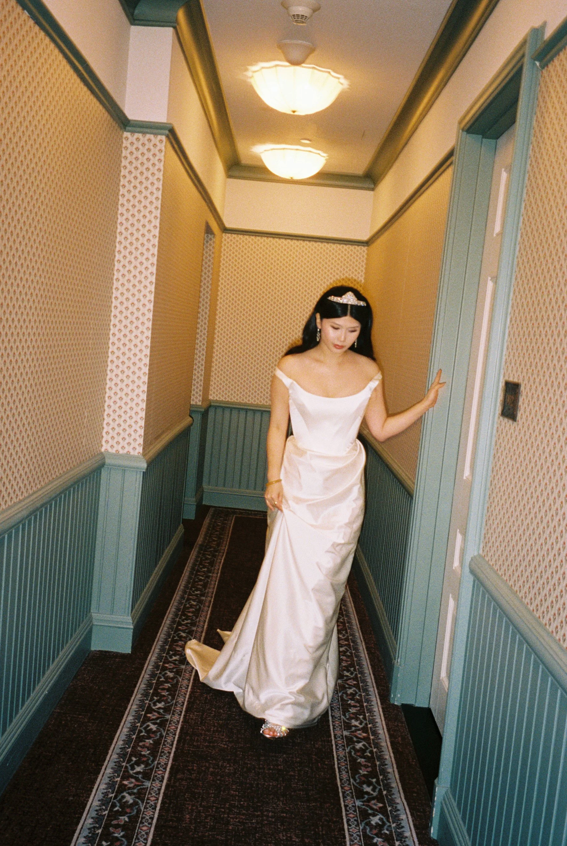 A woman in a white satin off-shoulder gown walking down a hallway with patterned wallpaper and a dark carpet runner. She is wearing a tiara and earrings, with her left hand touching the wall and her head slightly bowed.