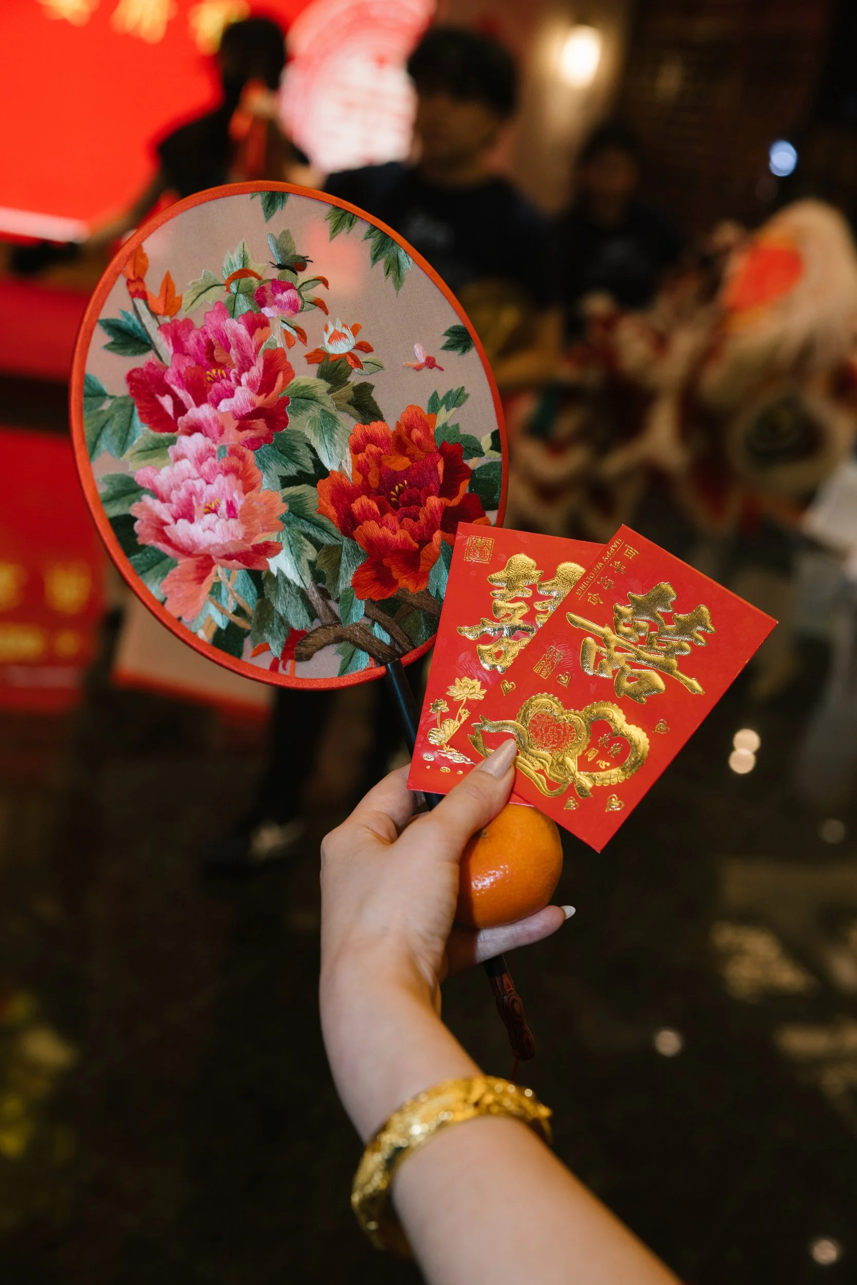 Person holding a traditional Chinese fan with floral design, red envelopes with gold Chinese characters, and a mandarin orange, celebrating a cultural event, with blurred people in the background.