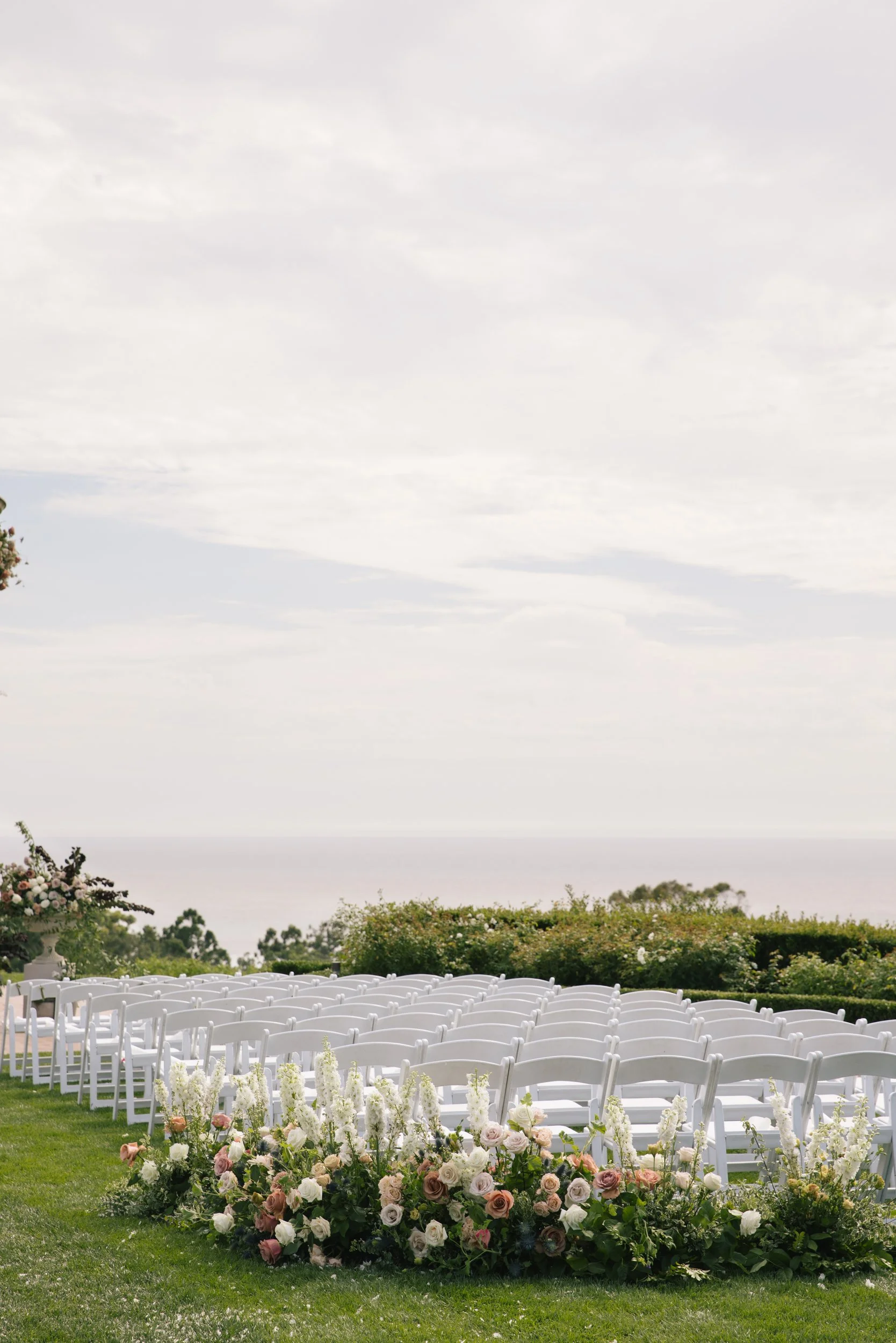 Outdoor wedding ceremony setup with white chairs arranged in rows, decorated with floral arrangements, under a cloudy sky with trees and bushes in the background.