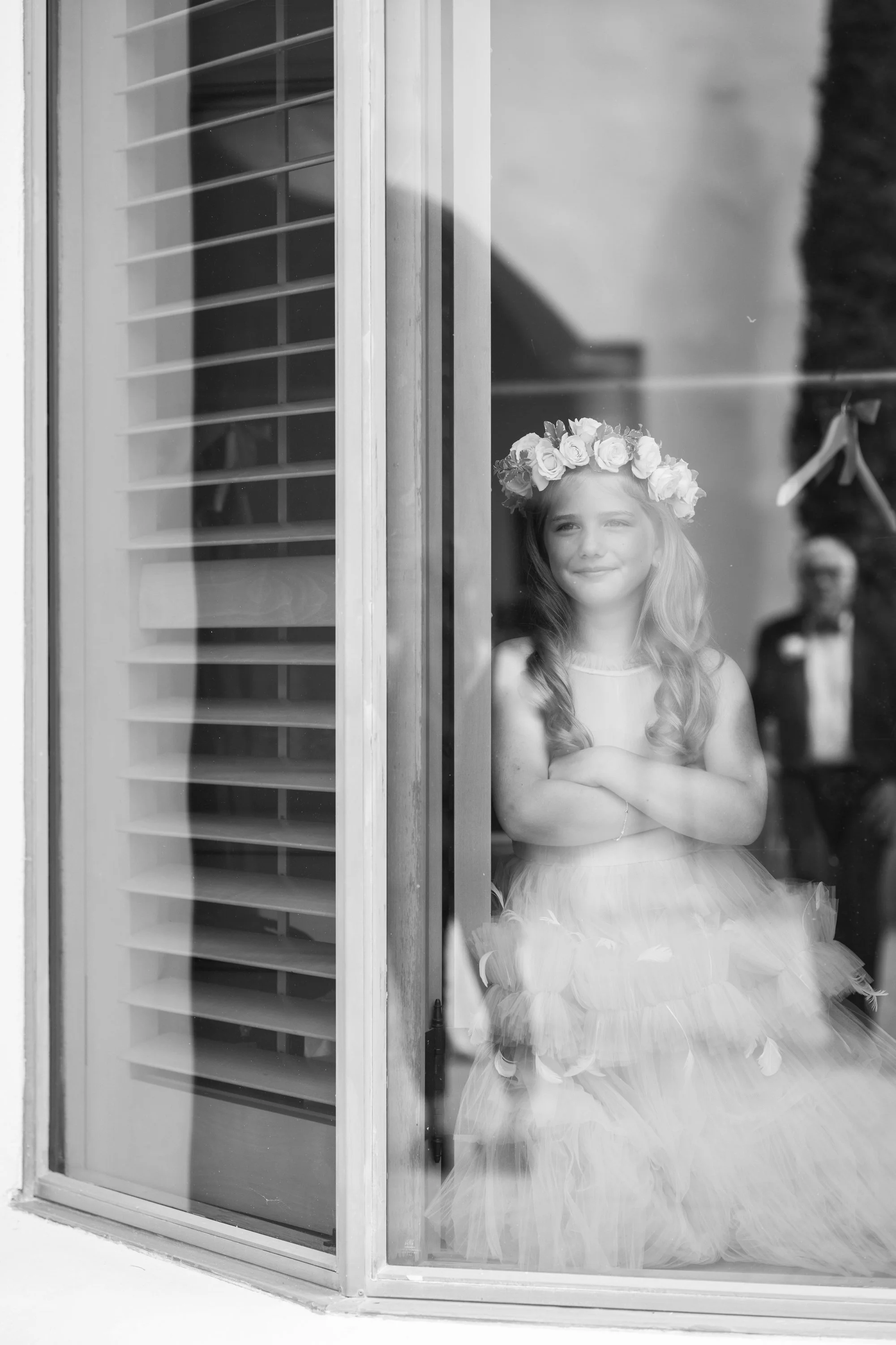 A young girl with a flower crown and a fluffy dress gazing through a glass window, with her arms crossed, and a man in a tuxedo visible outside the window in the background.