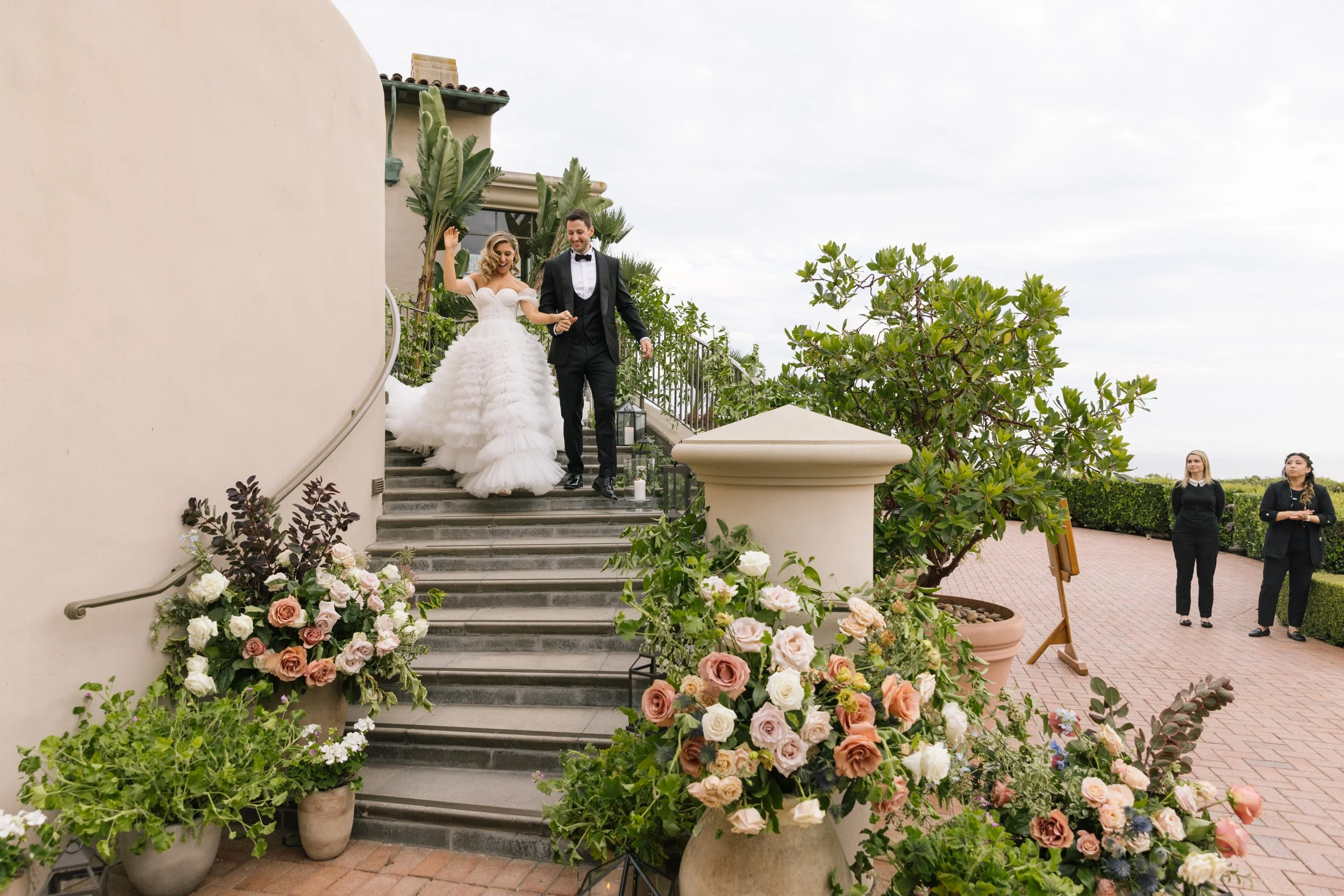 A bride and groom descending outdoor stairs decorated with pink, white, and peach flowers during a wedding ceremony, with two women standing nearby.