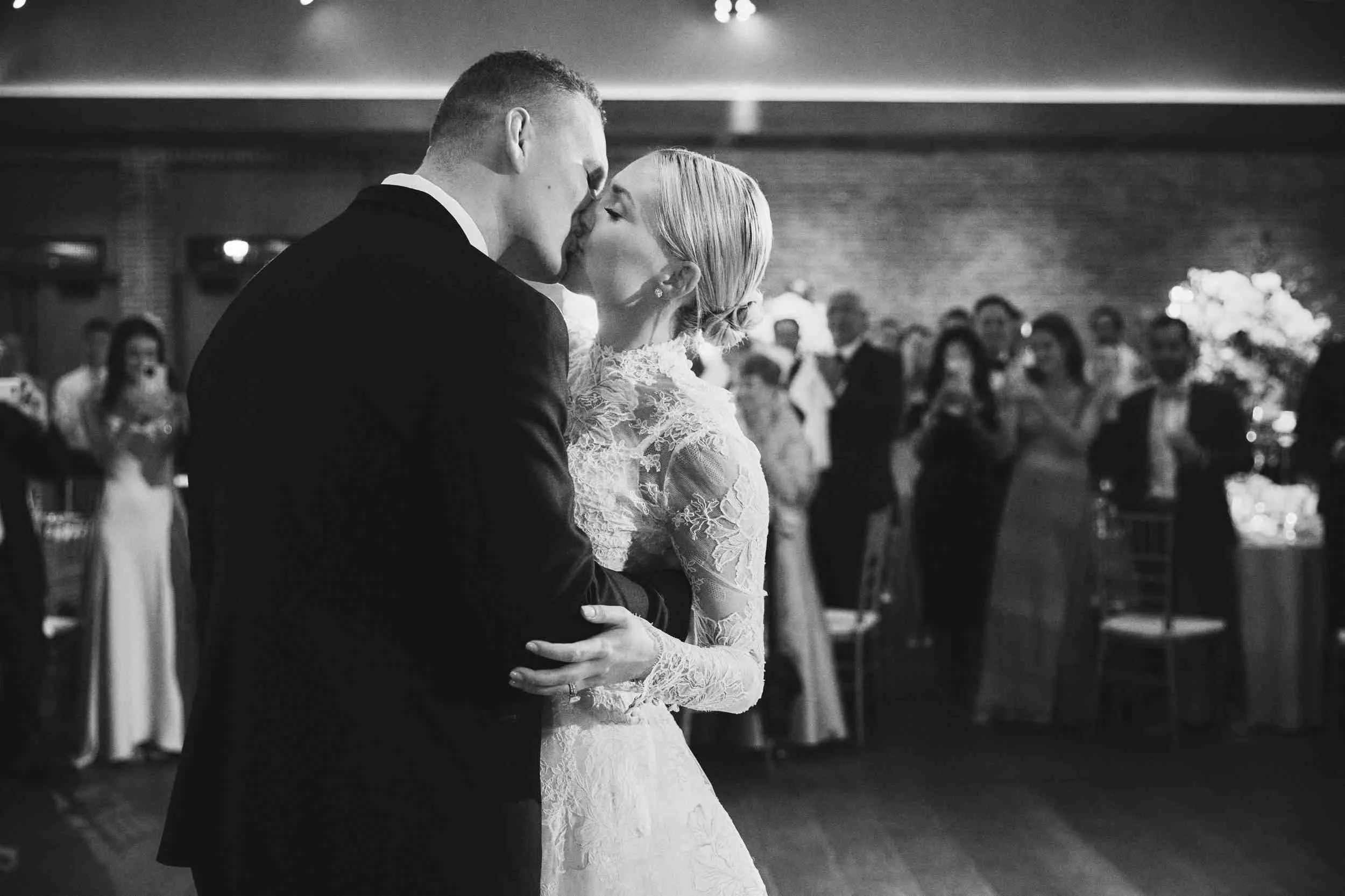 A black and white photo of a bride and groom dancing at their wedding reception, close-up of their faces and hands, with guests in the background.