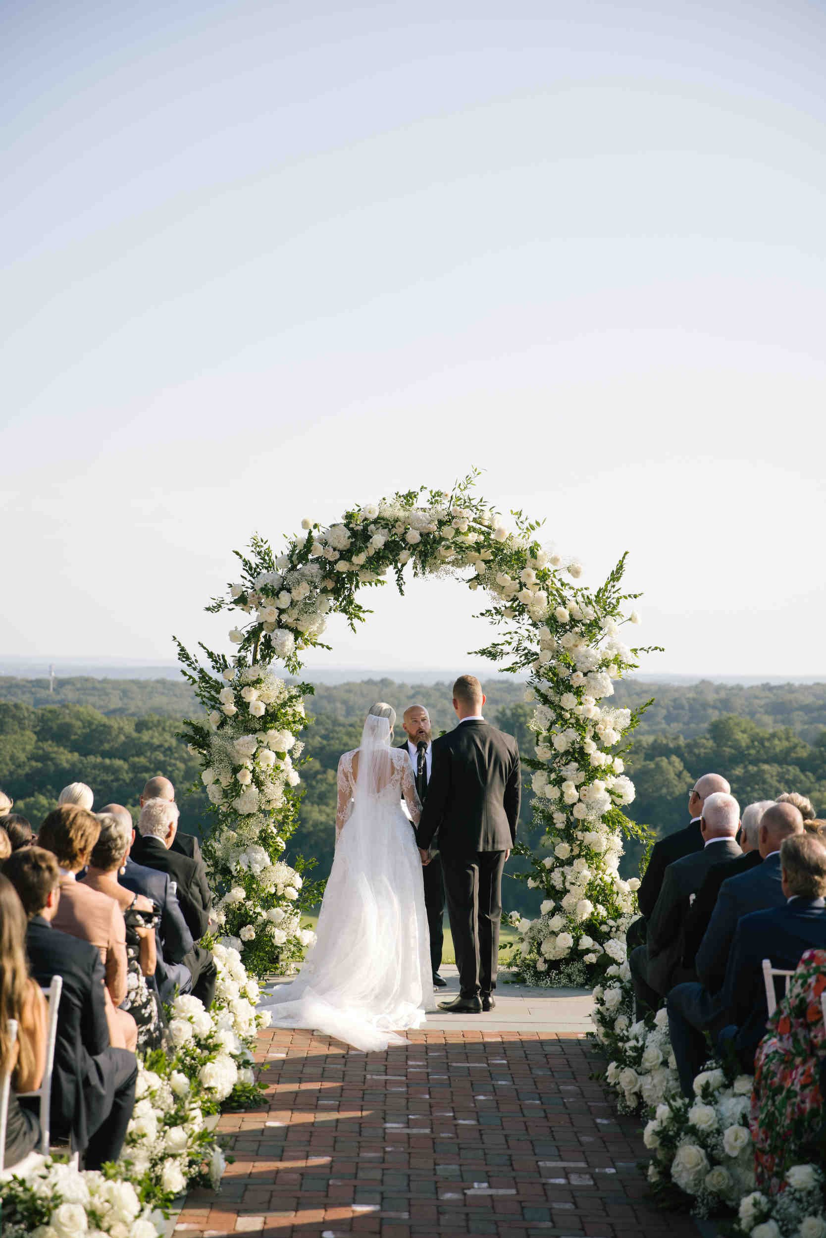 A wedding ceremony outdoors with a couple standing at an arched floral altar surrounded by guests. The bride is in a white wedding dress, the groom in a black suit. The ceremony has a scenic backdrop of green fields and trees.