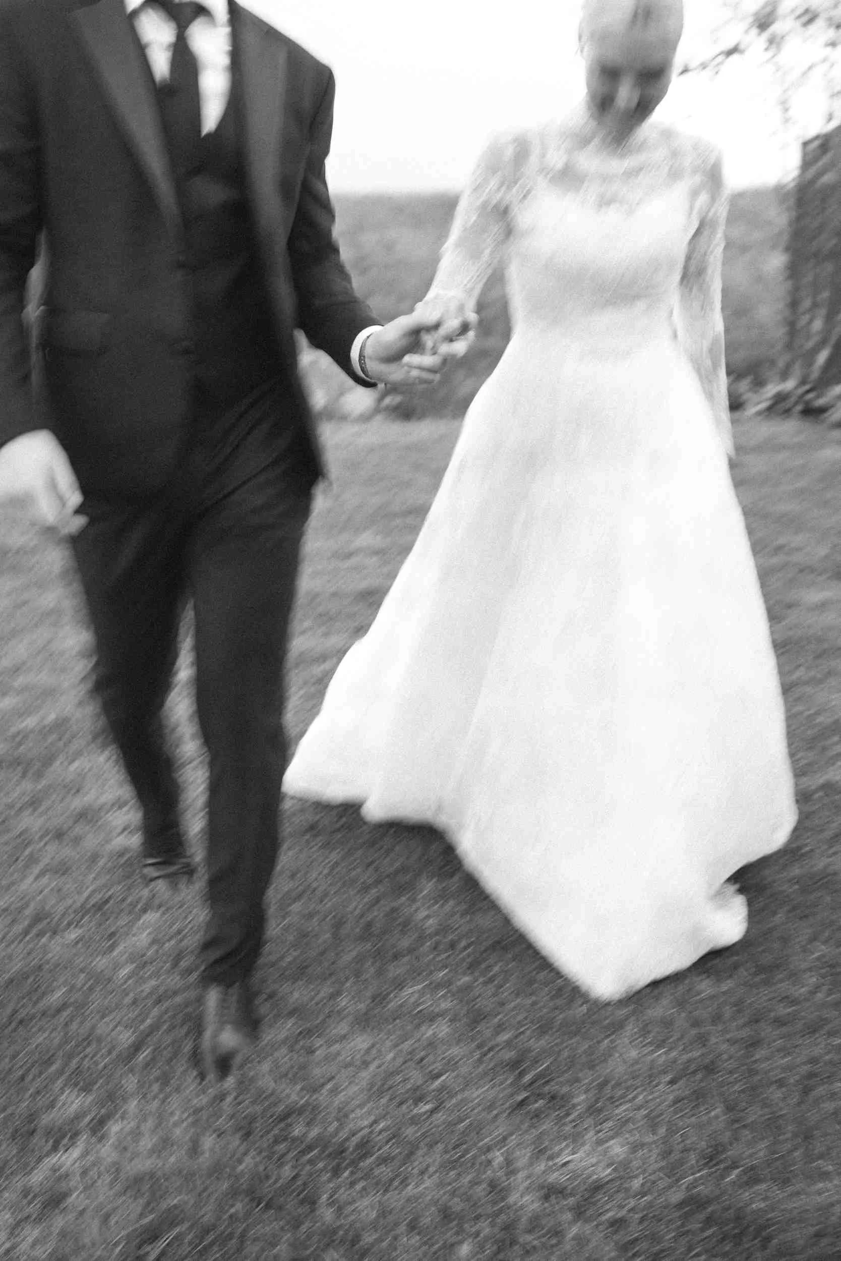 A black and white photo of a bride and groom holding hands, walking outdoors on grass. The groom is wearing a black suit with a tie, and the bride is wearing a long, lace wedding gown.