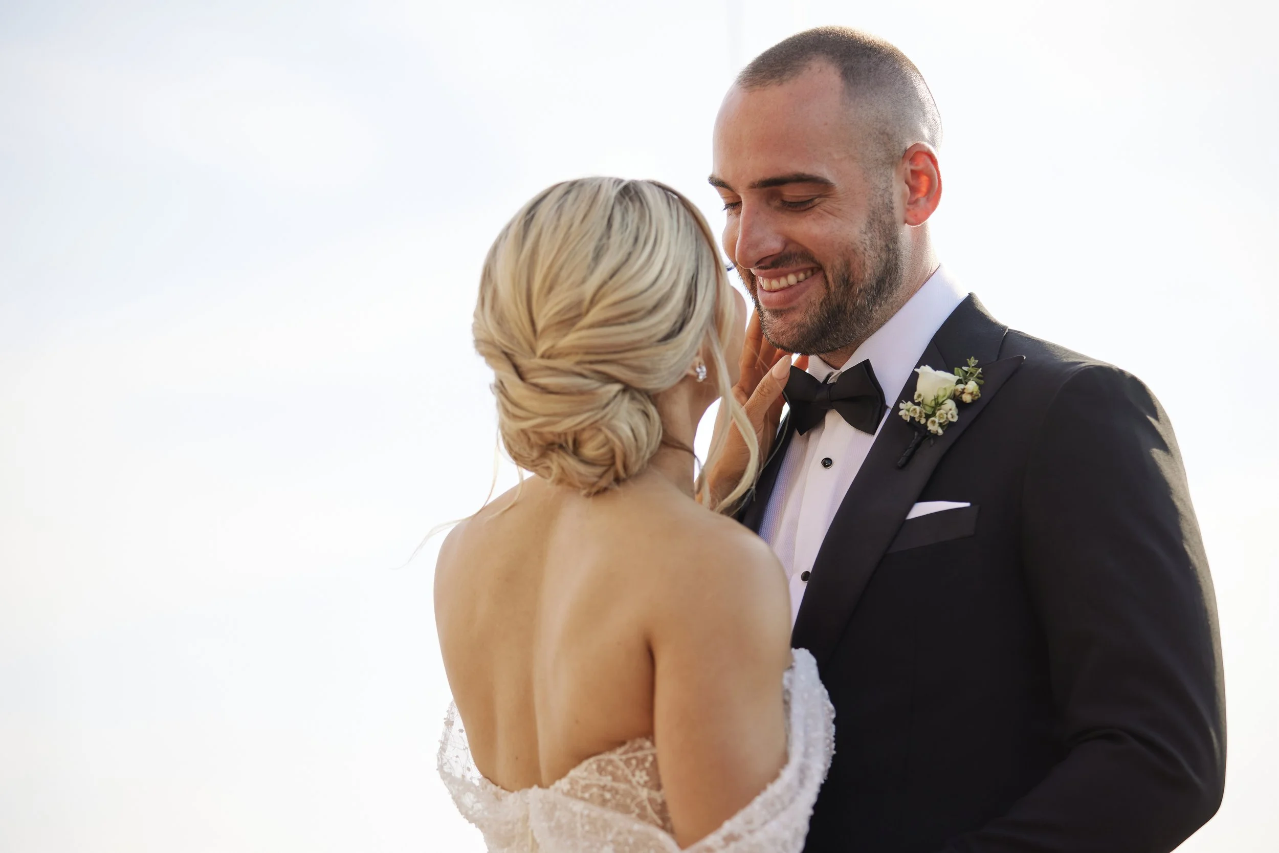 A bride and groom share an intimate moment outdoors on their wedding day. The bride is facing away from the camera with her back exposed, and the groom is smiling down at her, dressed in a tuxedo with a boutonnière. The background is bright and minim