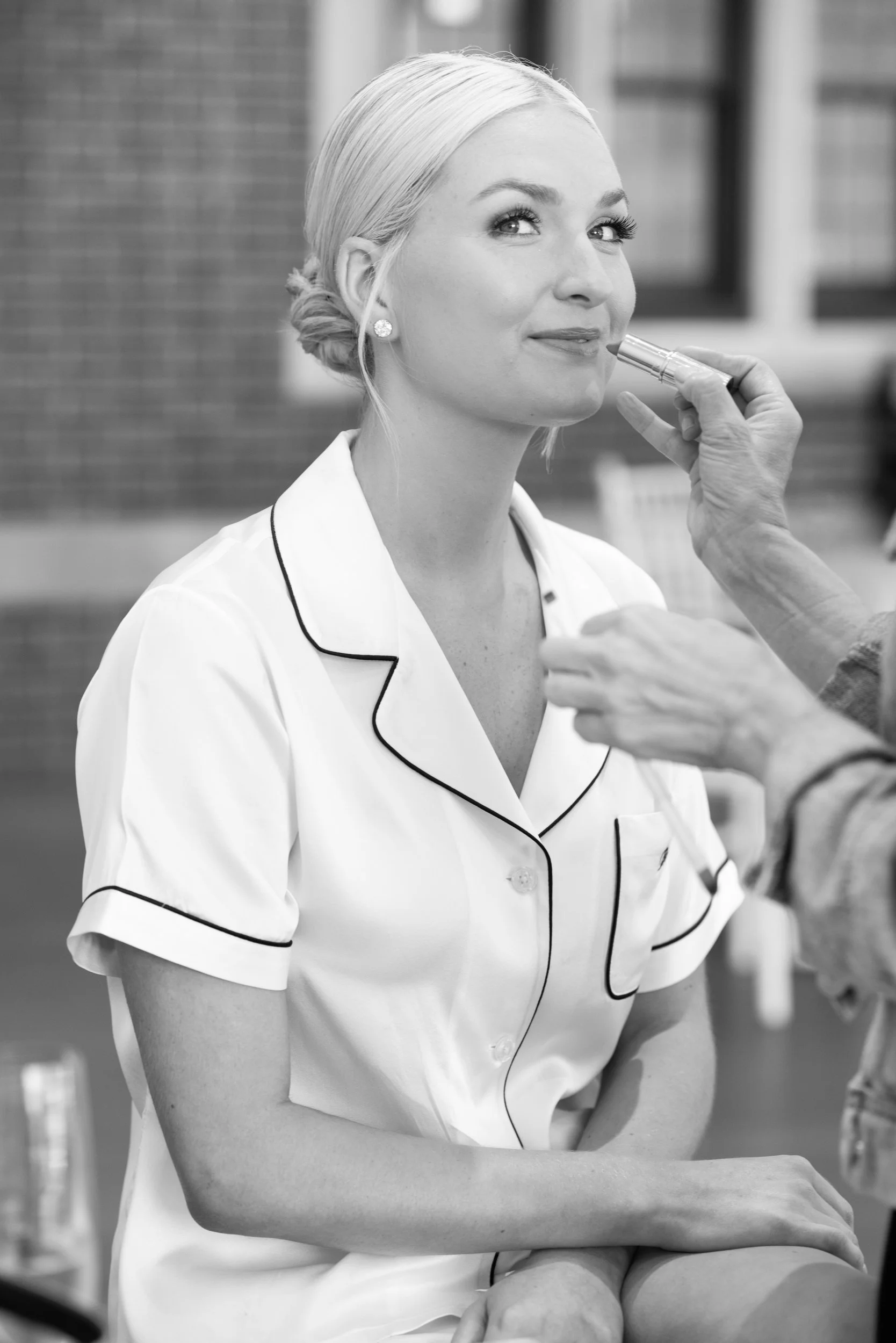 A woman with light hair styled in a bun, wearing pajamas, is getting lipstick applied by a person. She is looking slightly upward and smiling. The setting appears to be outdoors or in a casual environment.