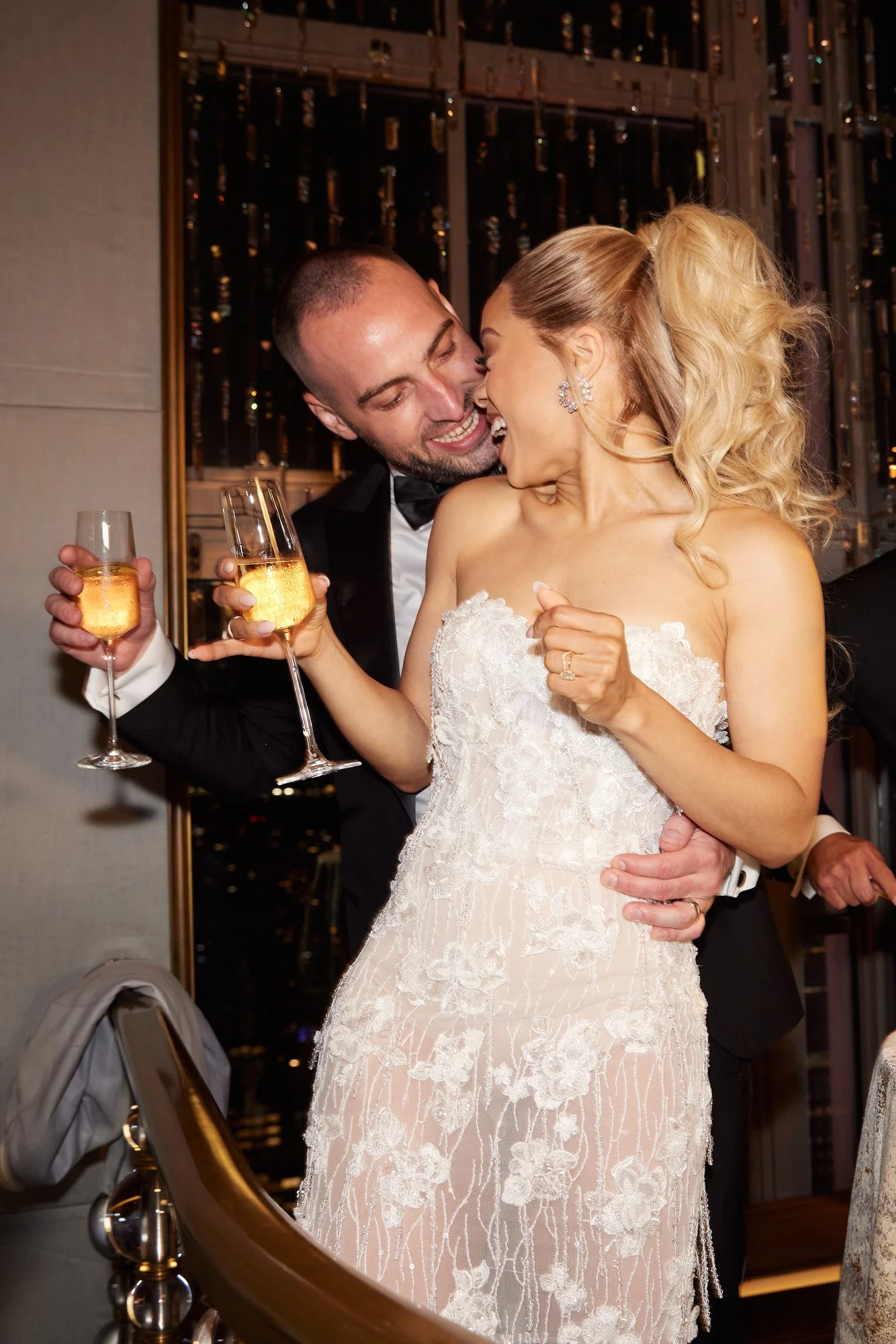 A newlywed couple celebrating with champagne at their wedding reception. The bride is wearing a white lace wedding gown with her hair styled in loose curls. The groom is dressed in a black tuxedo. They are smiling and leaning close to each other, hol
