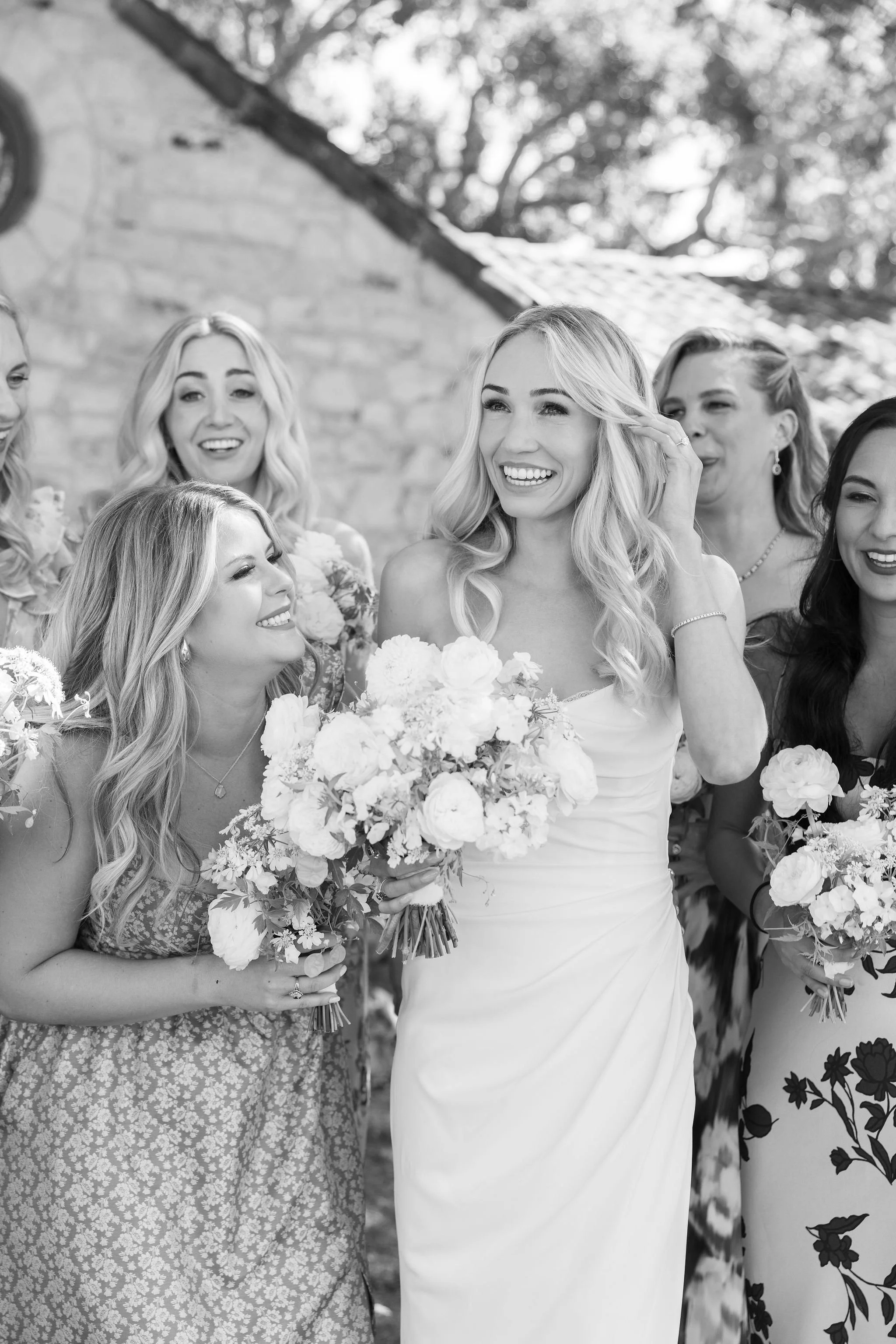 A black-and-white photo of a bride holding a bouquet of flowers, surrounded by bridesmaids, smiling and enjoying a moment outdoors.