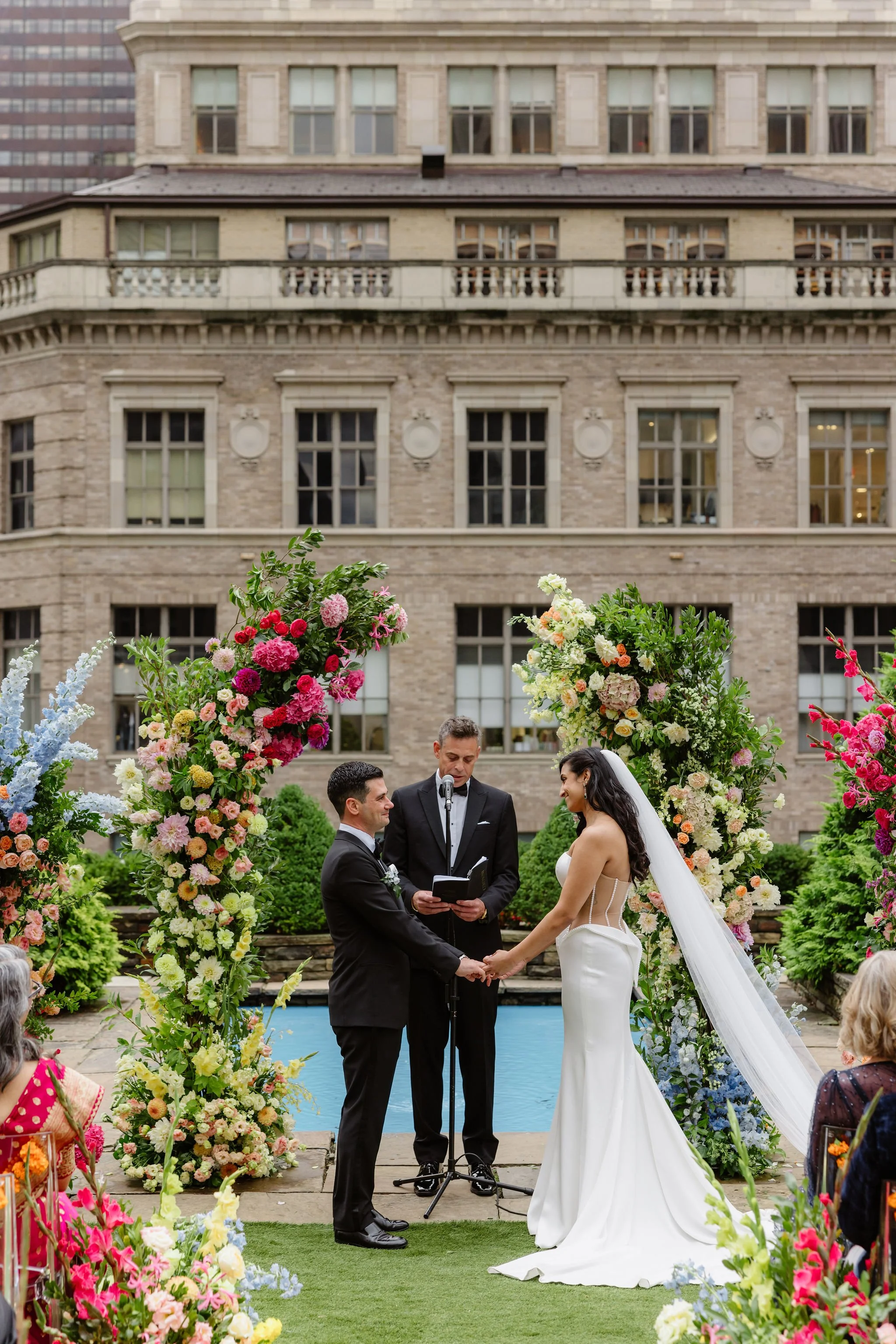A wedding ceremony outdoors with a bride and groom holding hands in front of an officiant, surrounded by colorful flowers, with a historic building in the background.