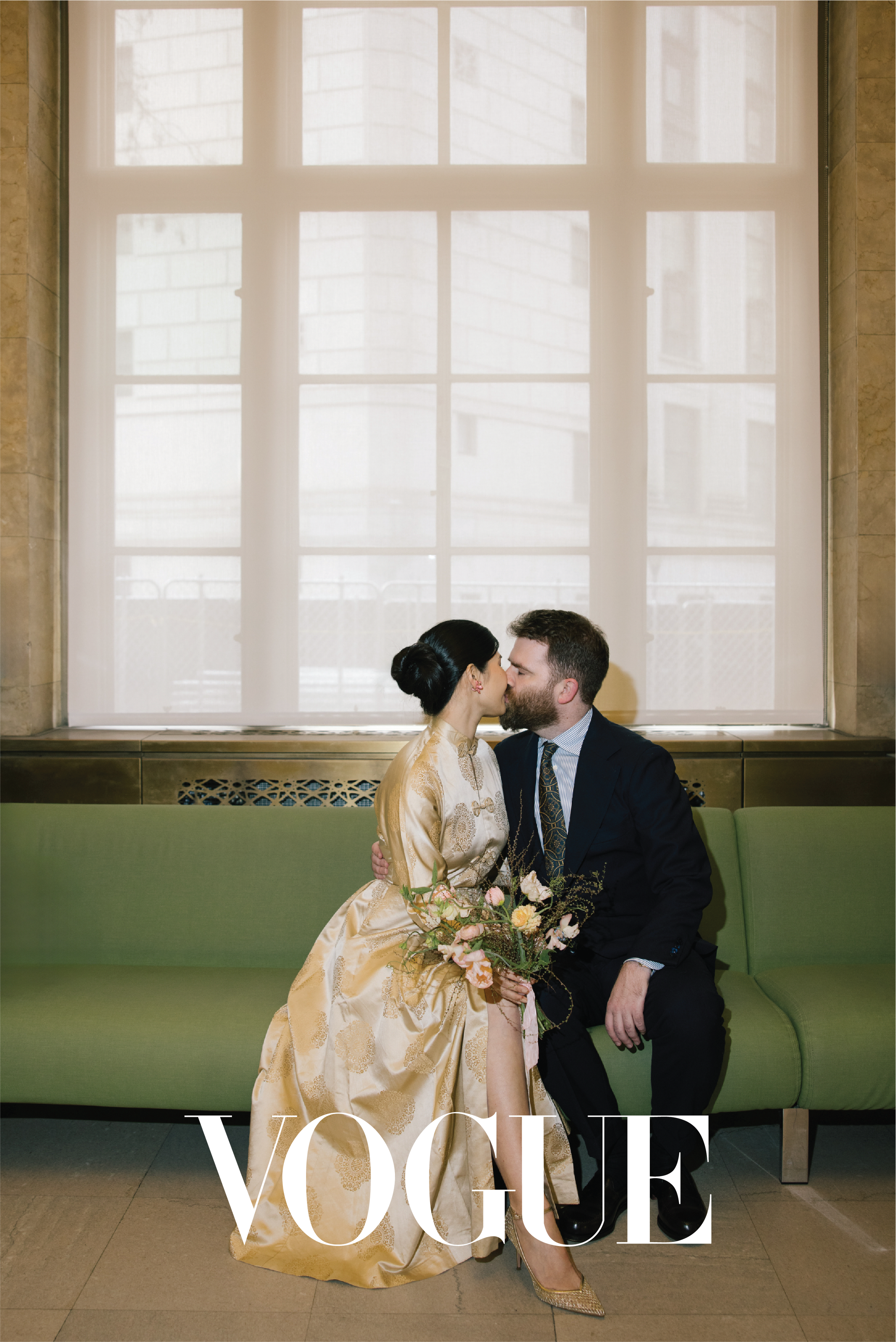 A couple in wedding attire sitting on a green bench, sharing a kiss inside a building with large window.