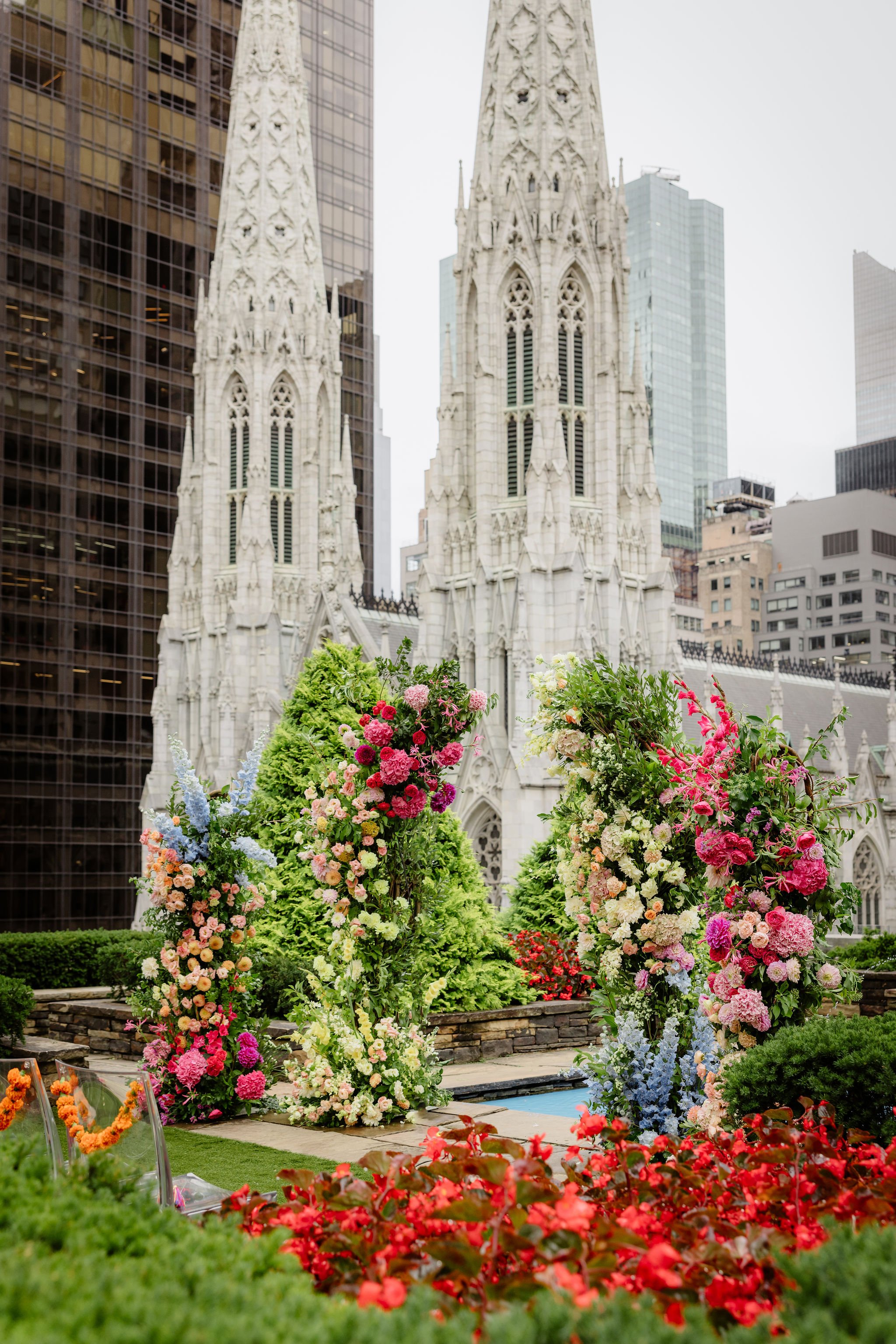 A garden with colorful flowers and green bushes in front of a gothic-style church with tall spires, surrounded by modern tall buildings.
