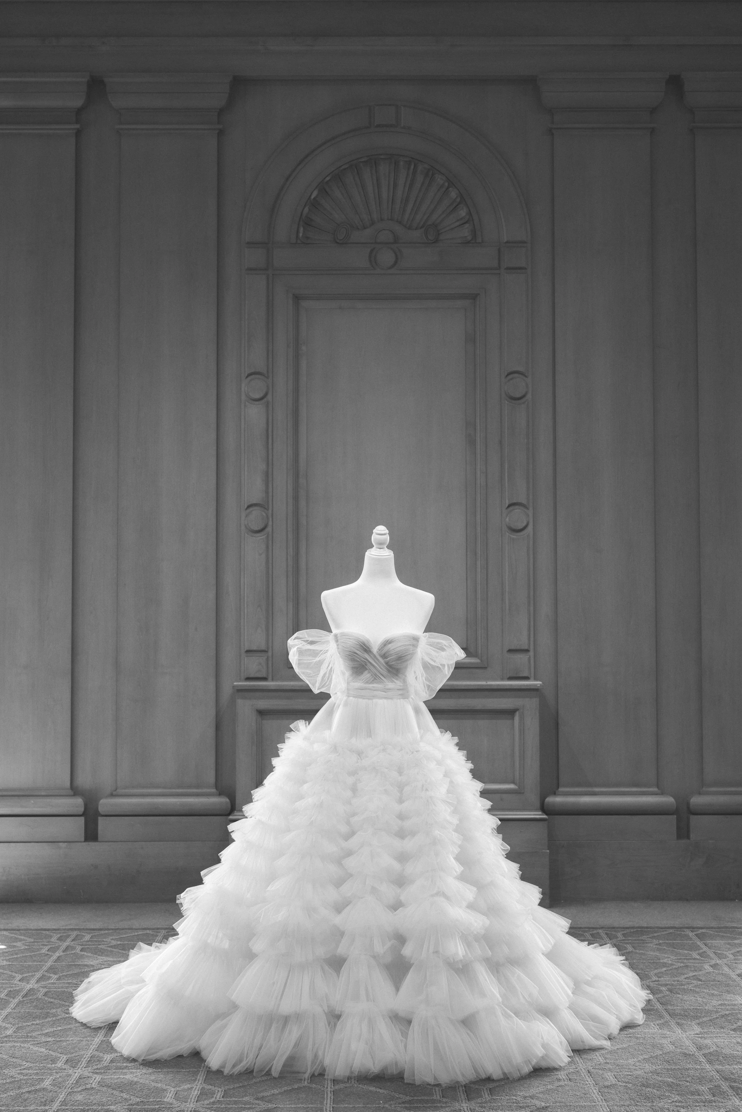 A white, ruffled wedding dress on a mannequin in a room with wood-paneled walls and a carpeted floor.