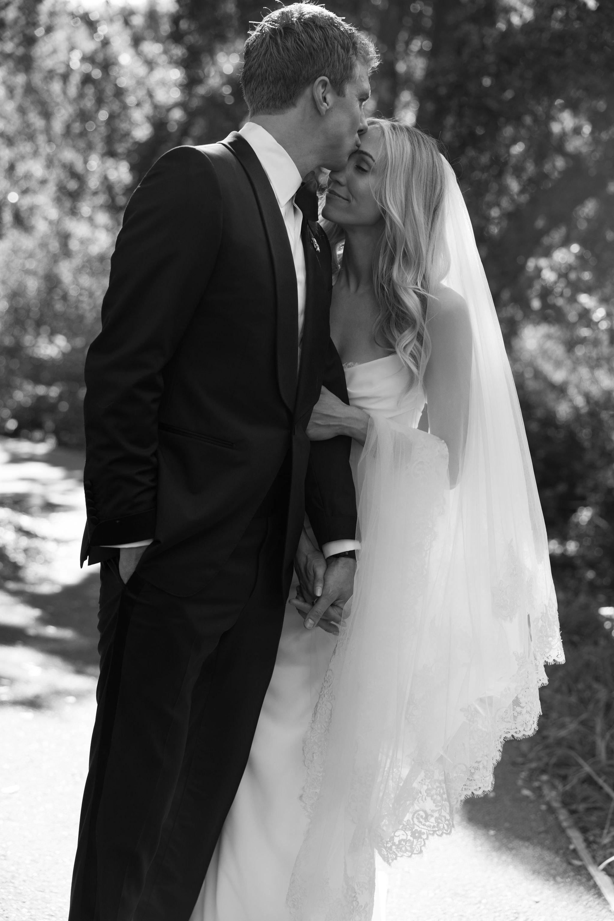 A black and white photograph of a newlywed couple outdoors, holding hands. The groom, in a tuxedo, kisses the bride on her forehead. The bride wears a white wedding dress with lace details and a veil. They are surrounded by blurred trees and foliage.