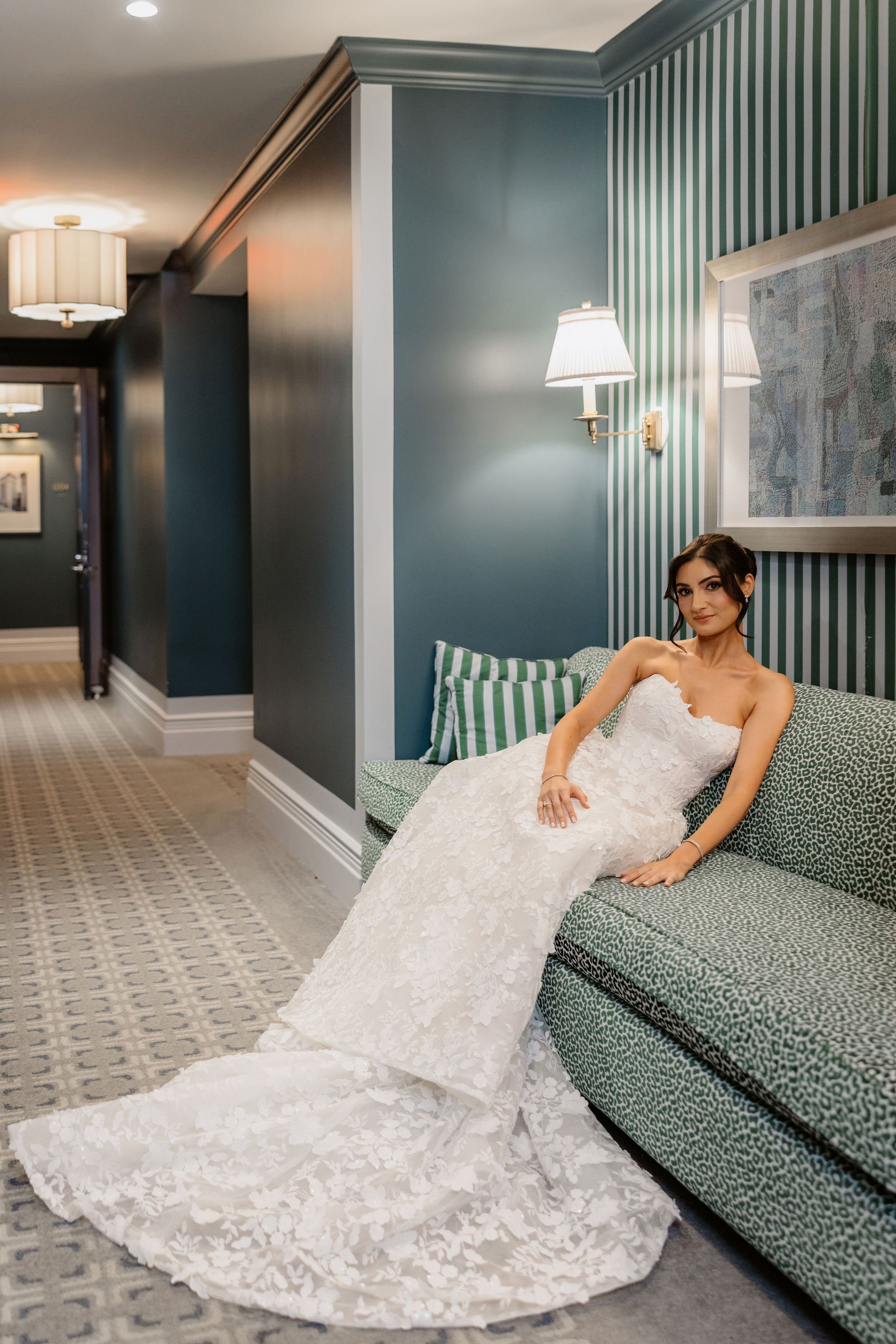 A woman in a white wedding dress sitting on a green patterned couch in a hotel room with striped wallpaper and wall lamps.