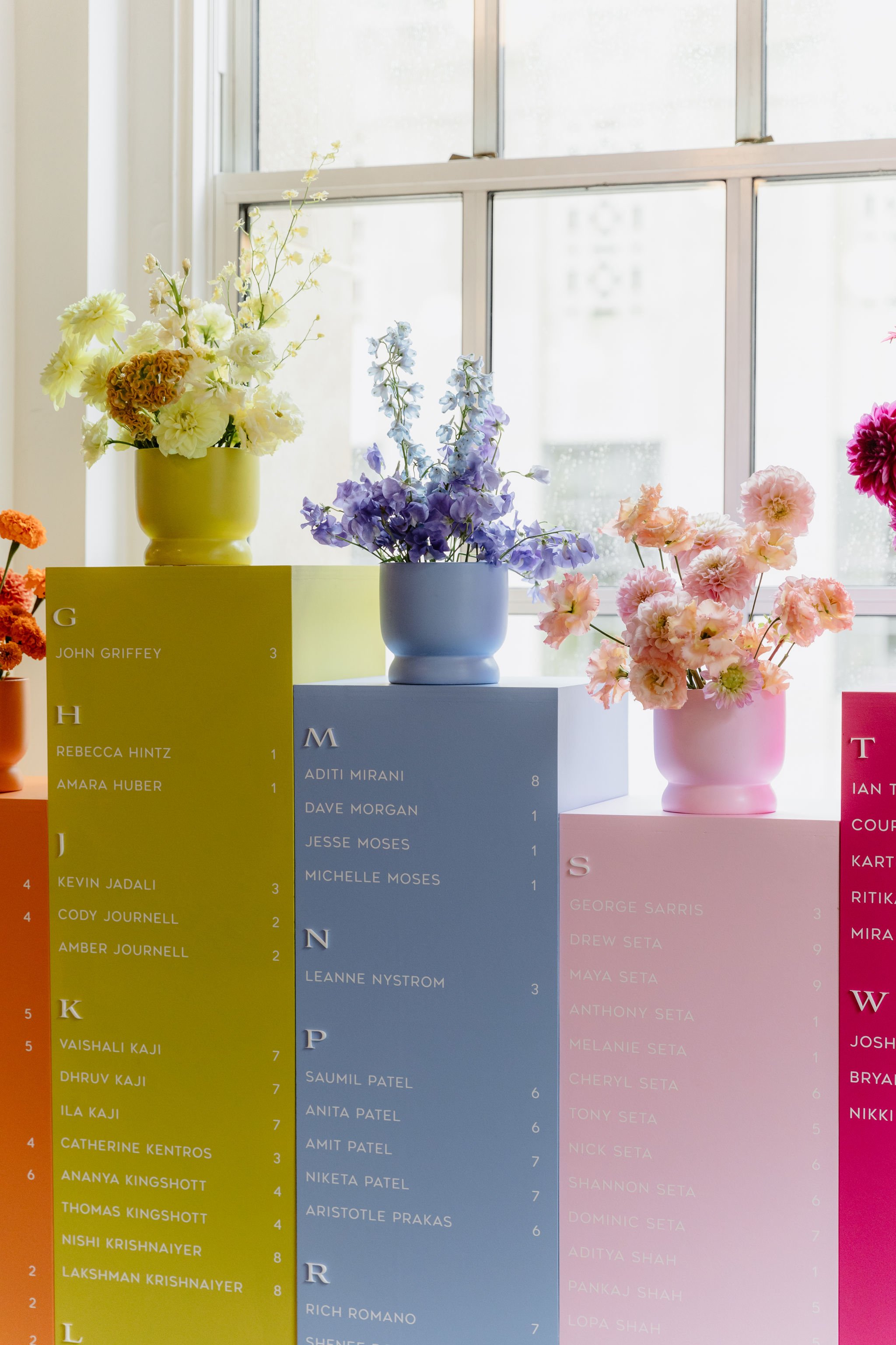 Colorful flower arrangements in vases on display, with name tags and rankings from a flower competition, set against a windowed background.