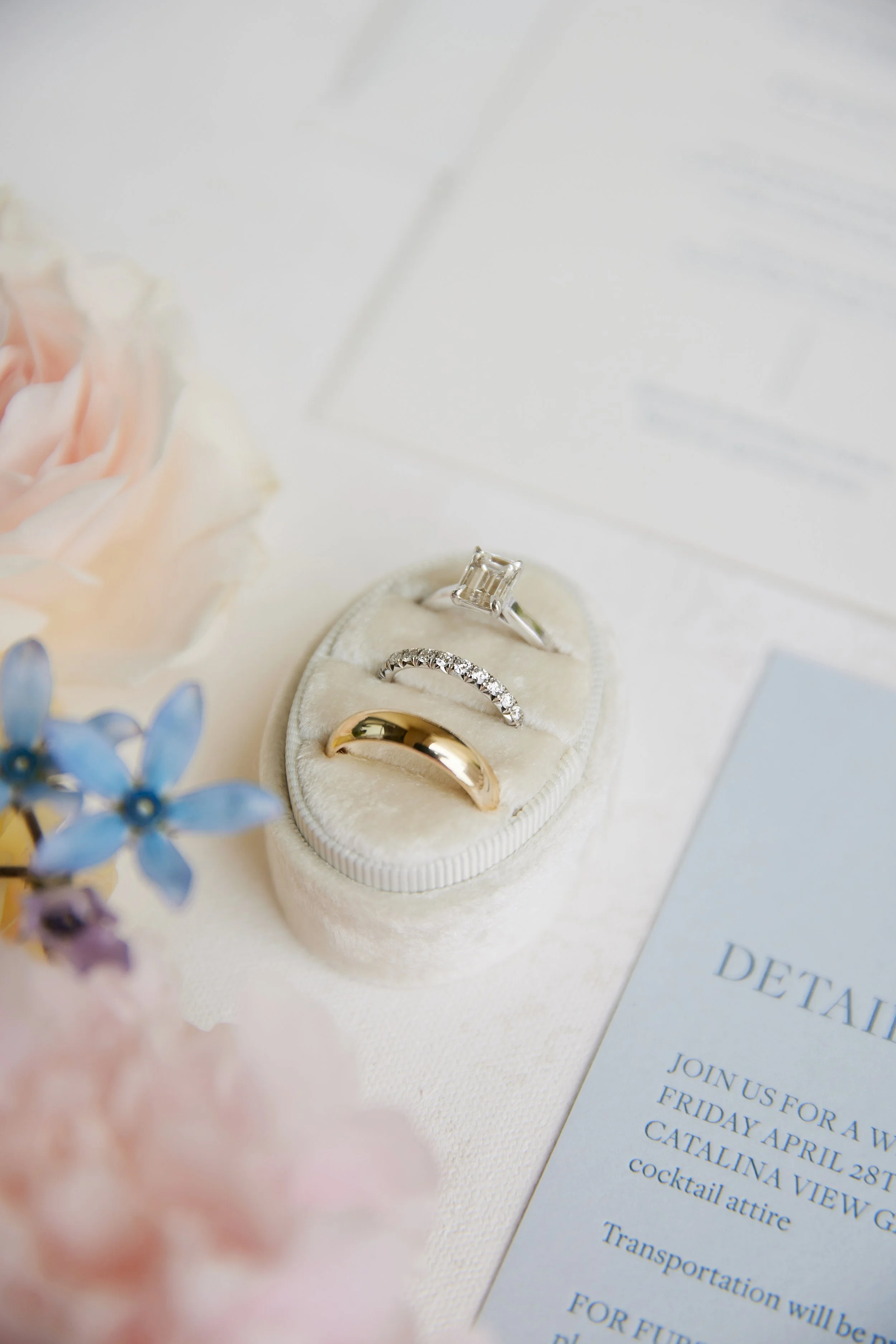 Close-up of a jewelry box containing three rings, with a blurred pink flower and a blue flower in the foreground, and a printed event invitation partially visible in the background.