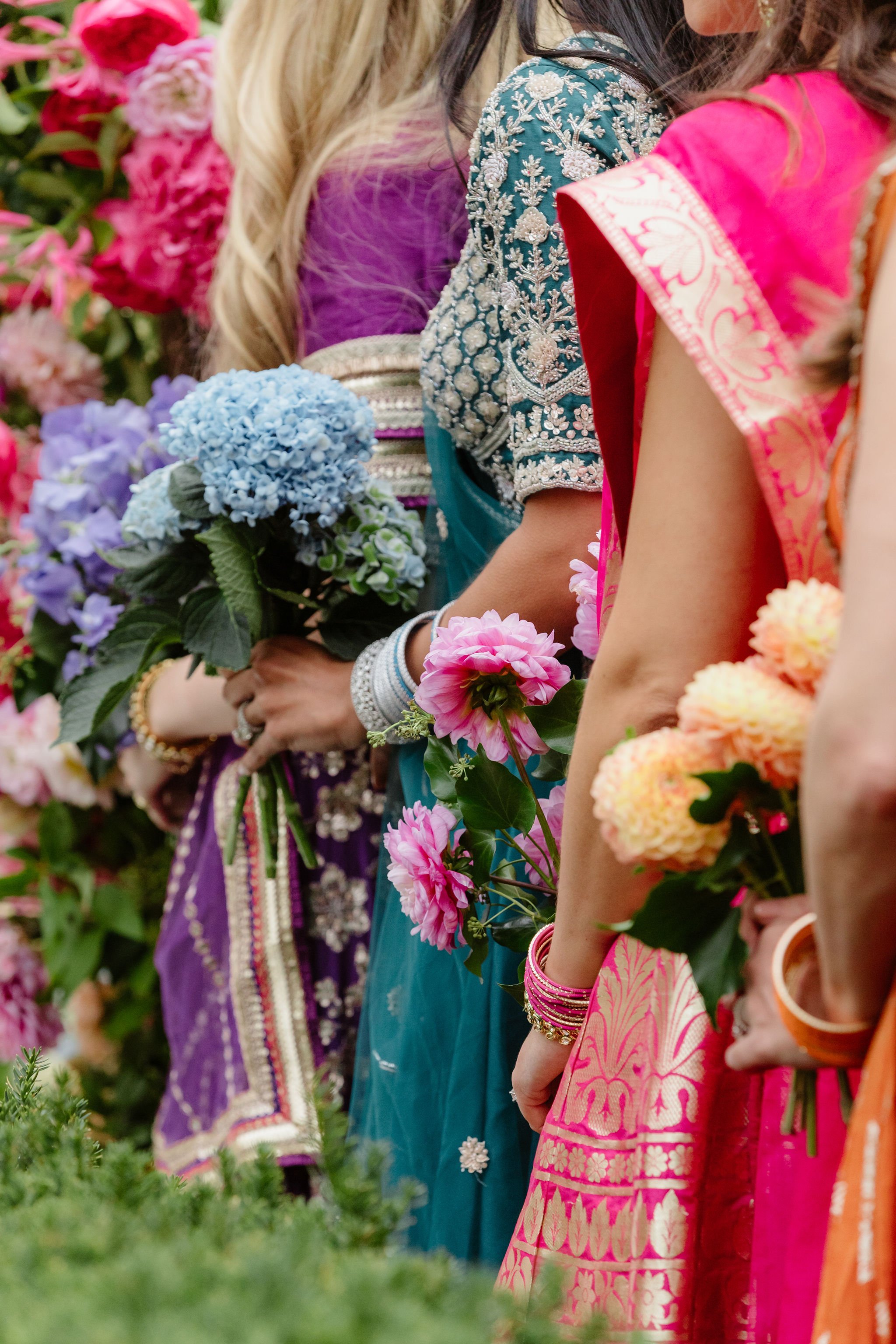 A line of women dressed in colorful traditional Indian sarees and adorned with jewelry holding bouquets of pink, purple, and blue flowers.