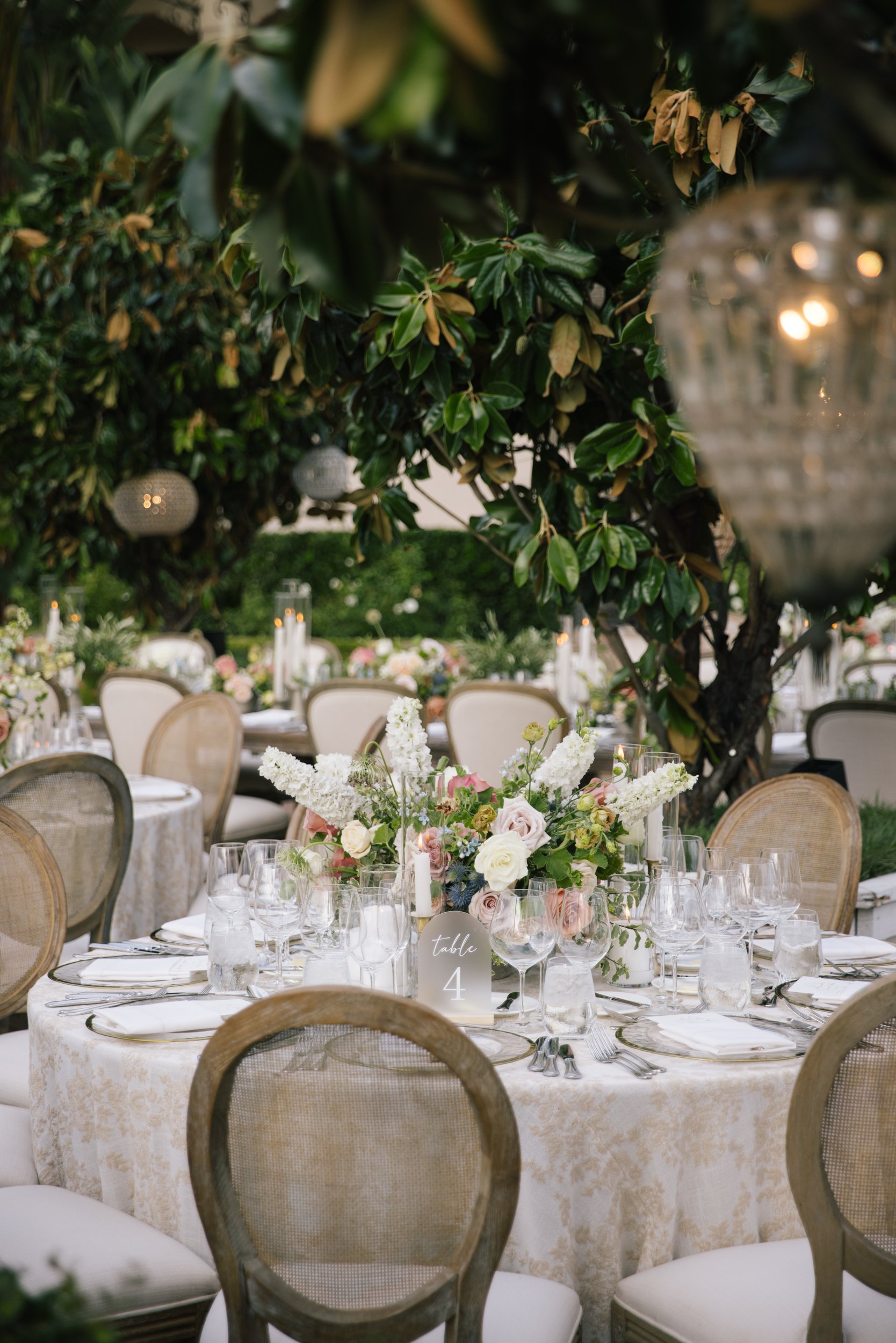 Elegant outdoor wedding reception table decorated with a floral centerpiece, white tablecloth, glassware, and table number 4, under green foliage and hanging lanterns.