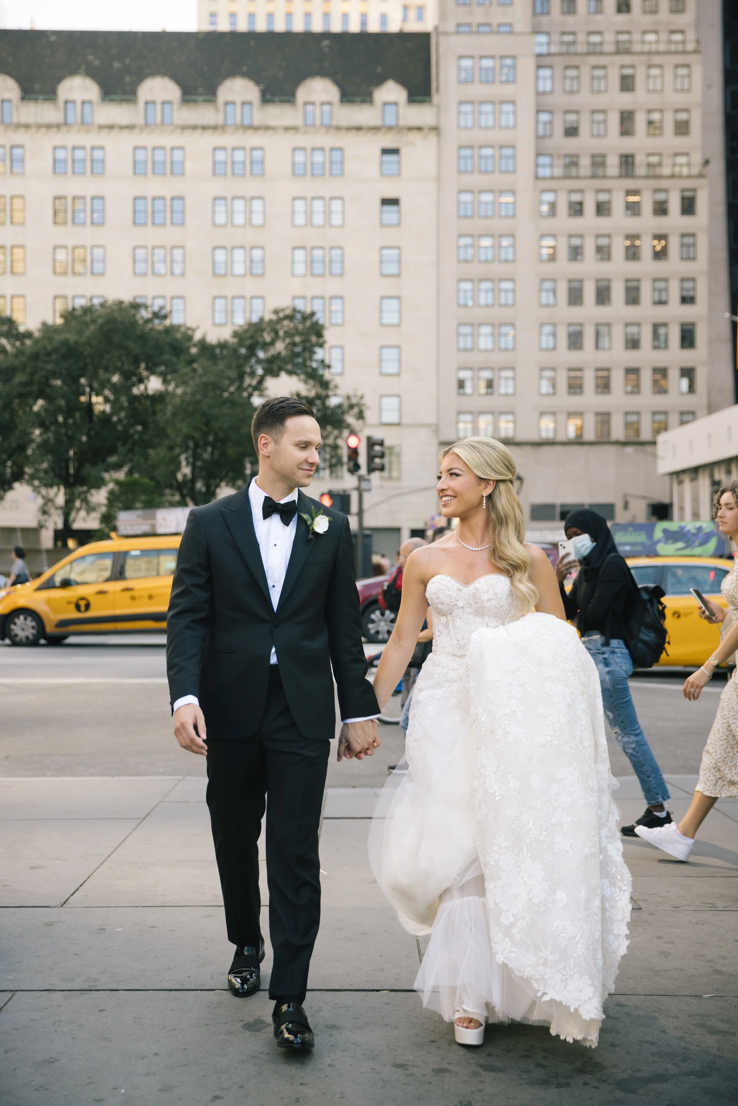 A bride and groom holding hands in a city street, surrounded by pedestrians and yellow taxis, with tall buildings in the background.