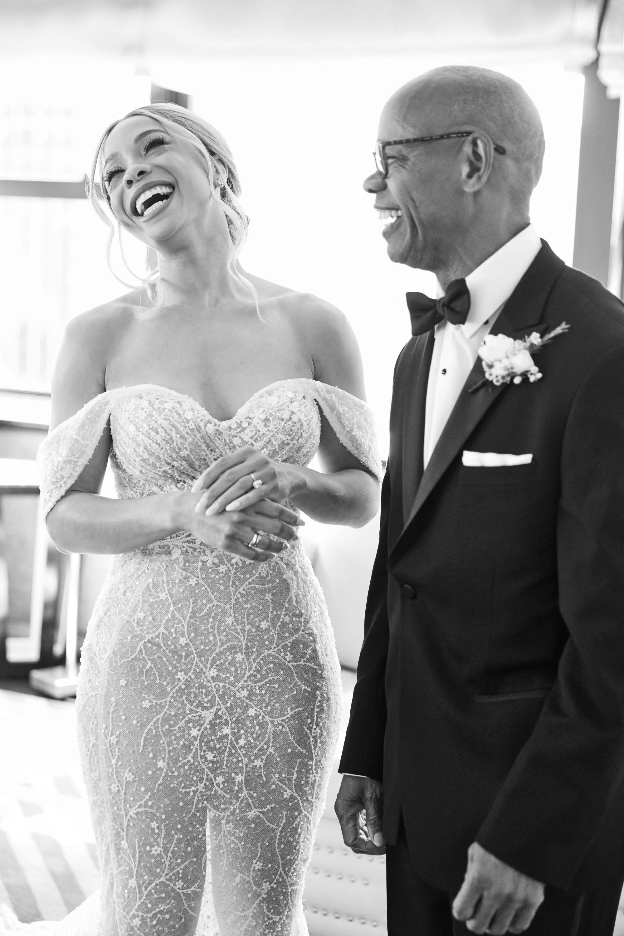 A bride in a lace wedding dress laughing and smiling while holding hands with a man dressed in a tuxedo, during a wedding ceremony.