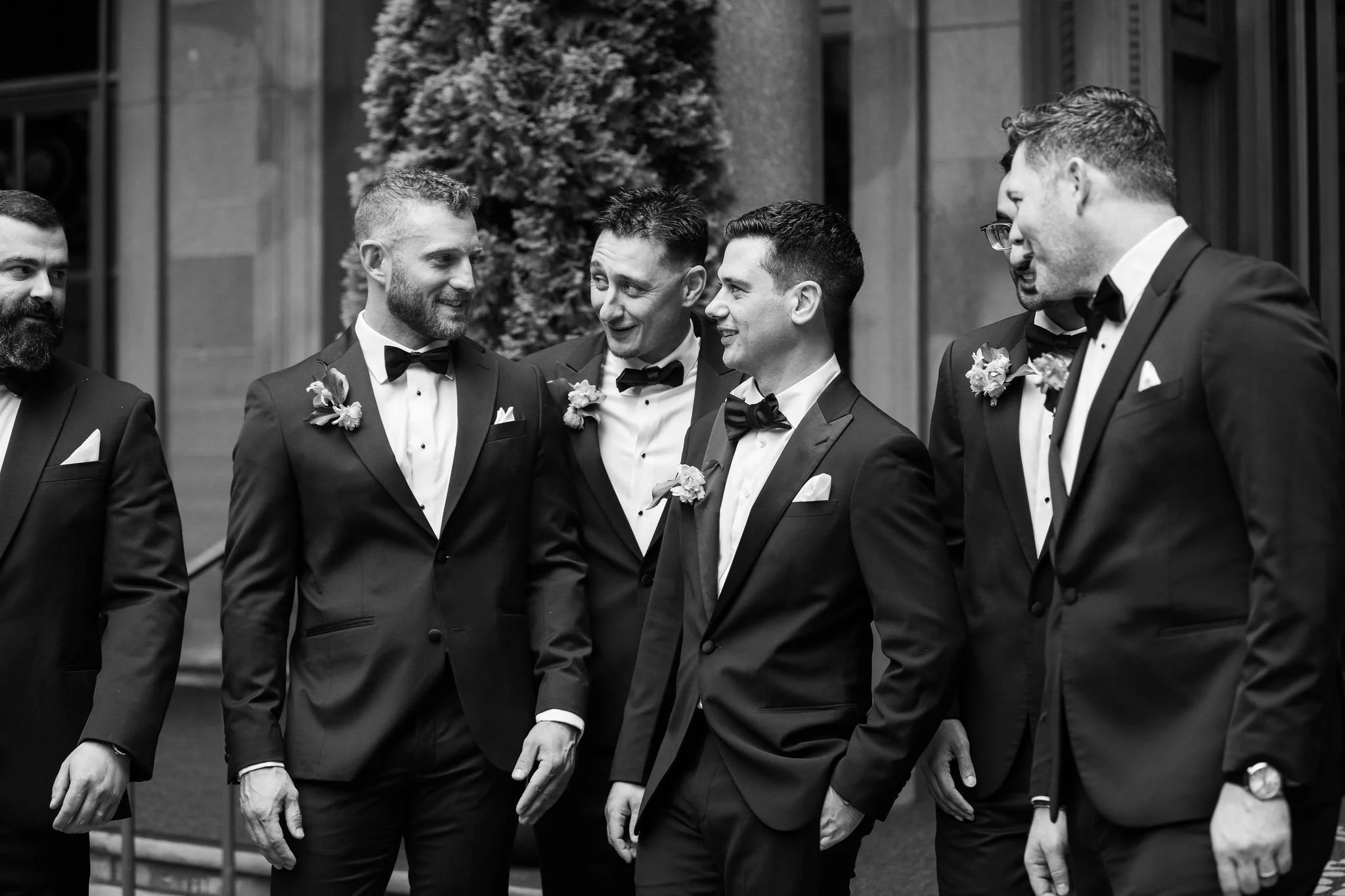 Group of men in tuxedos and bow ties, talking and smiling outside in front of a building with trees.