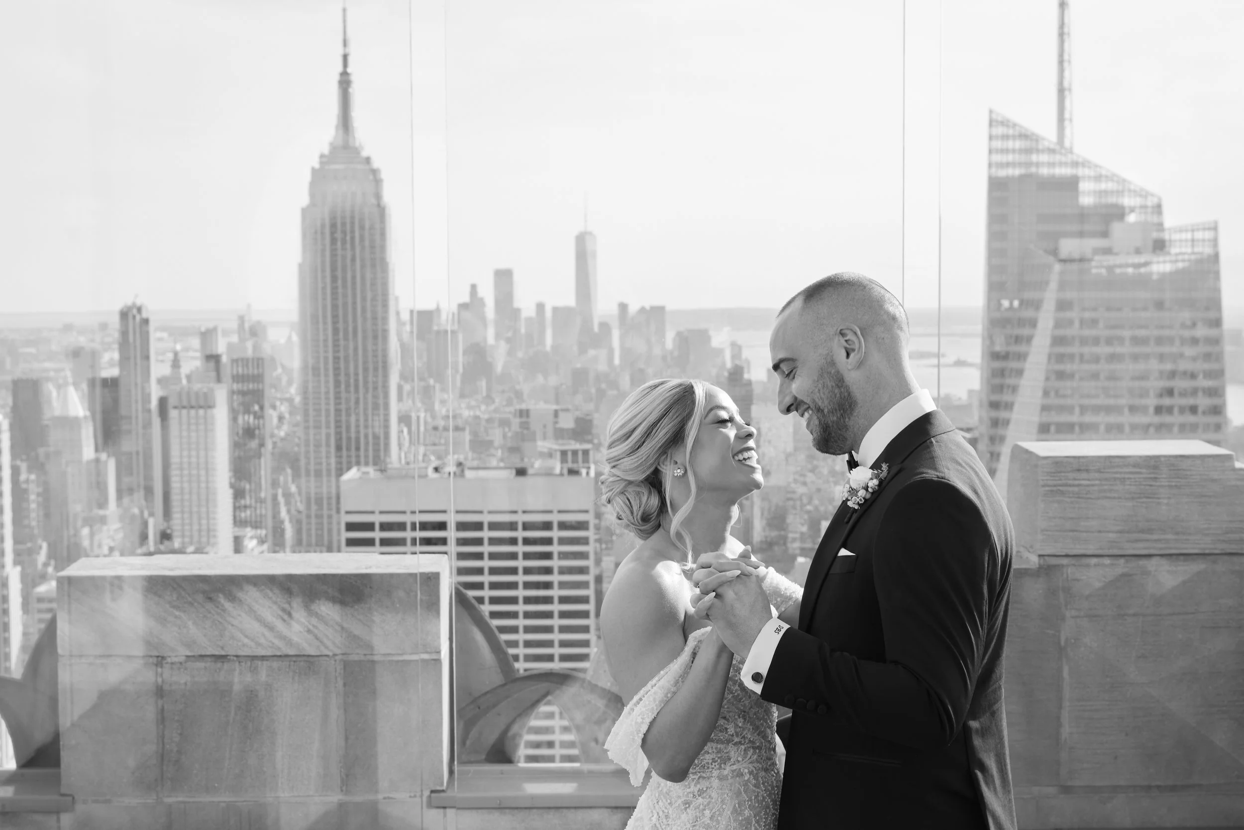 A bride and groom smiling and holding hands during their wedding on a rooftop with a view of the New York City skyline, including the Empire State Building and One World Trade Center.