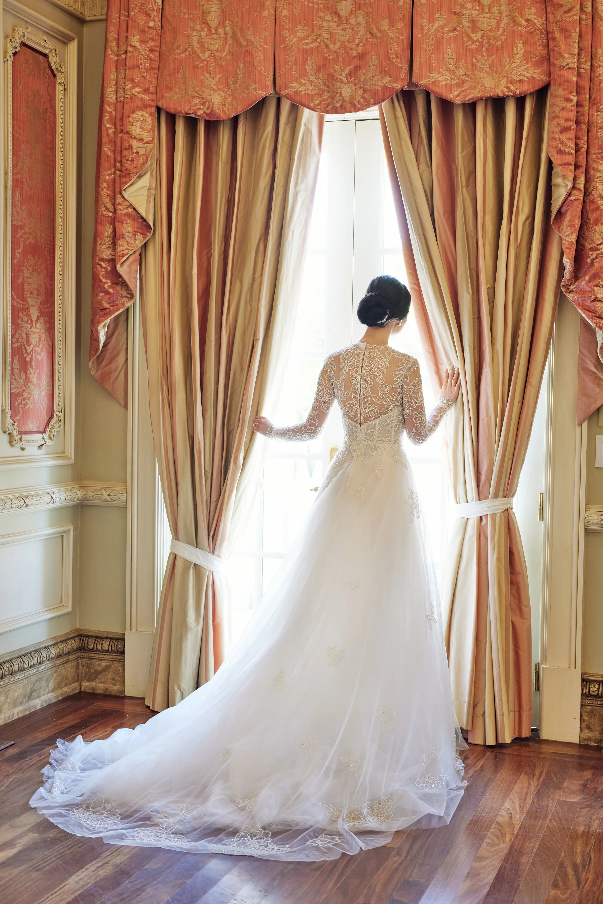 A bride in a lace wedding dress standing in front of a large window, holding back peach and cream curtains with floral patterns, with her back to the camera.