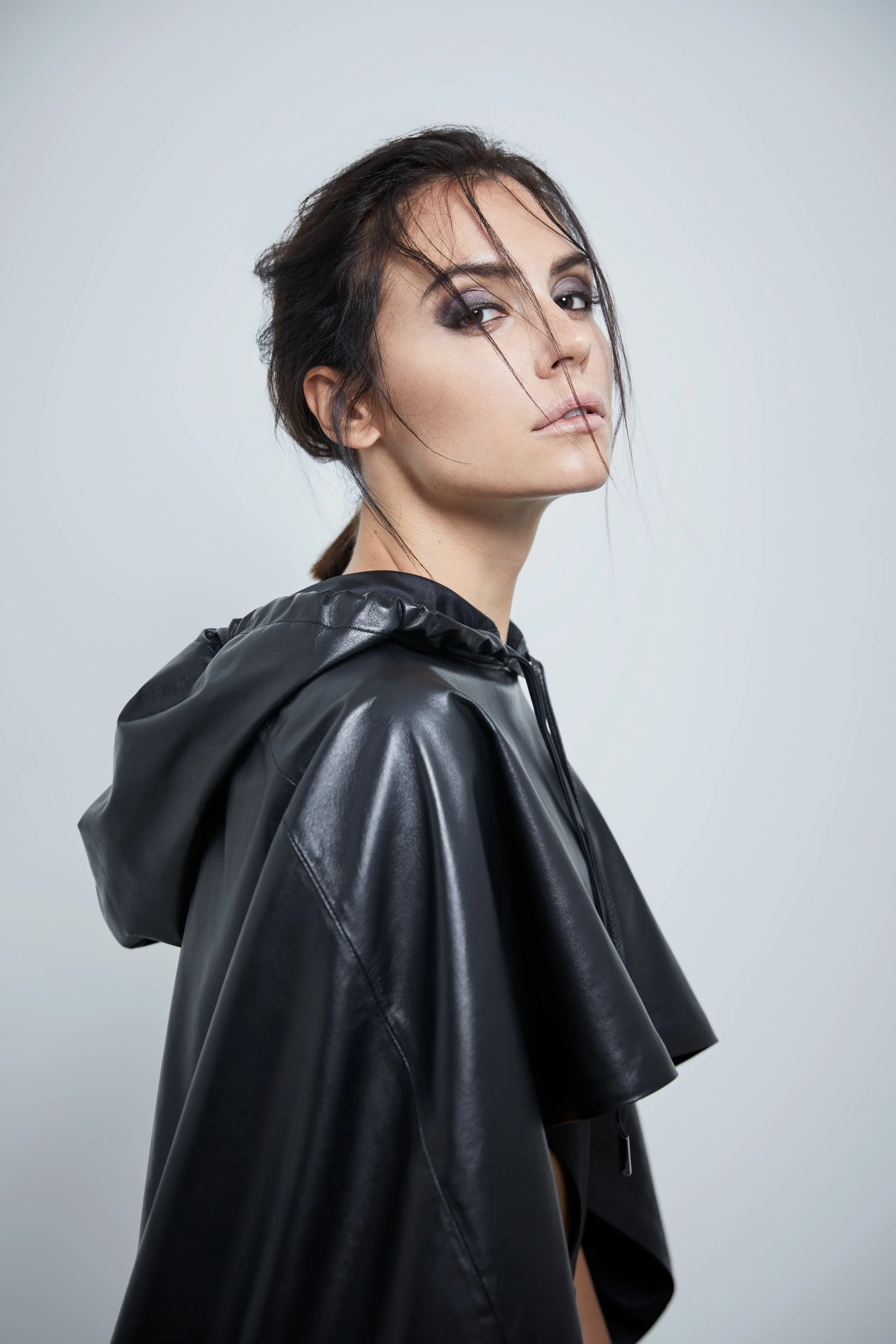 Portrait of a woman with dark hair styled in a messy bun, wearing a black hooded leather jacket, with a neutral expression and slightly tousled hair, against a plain light background.