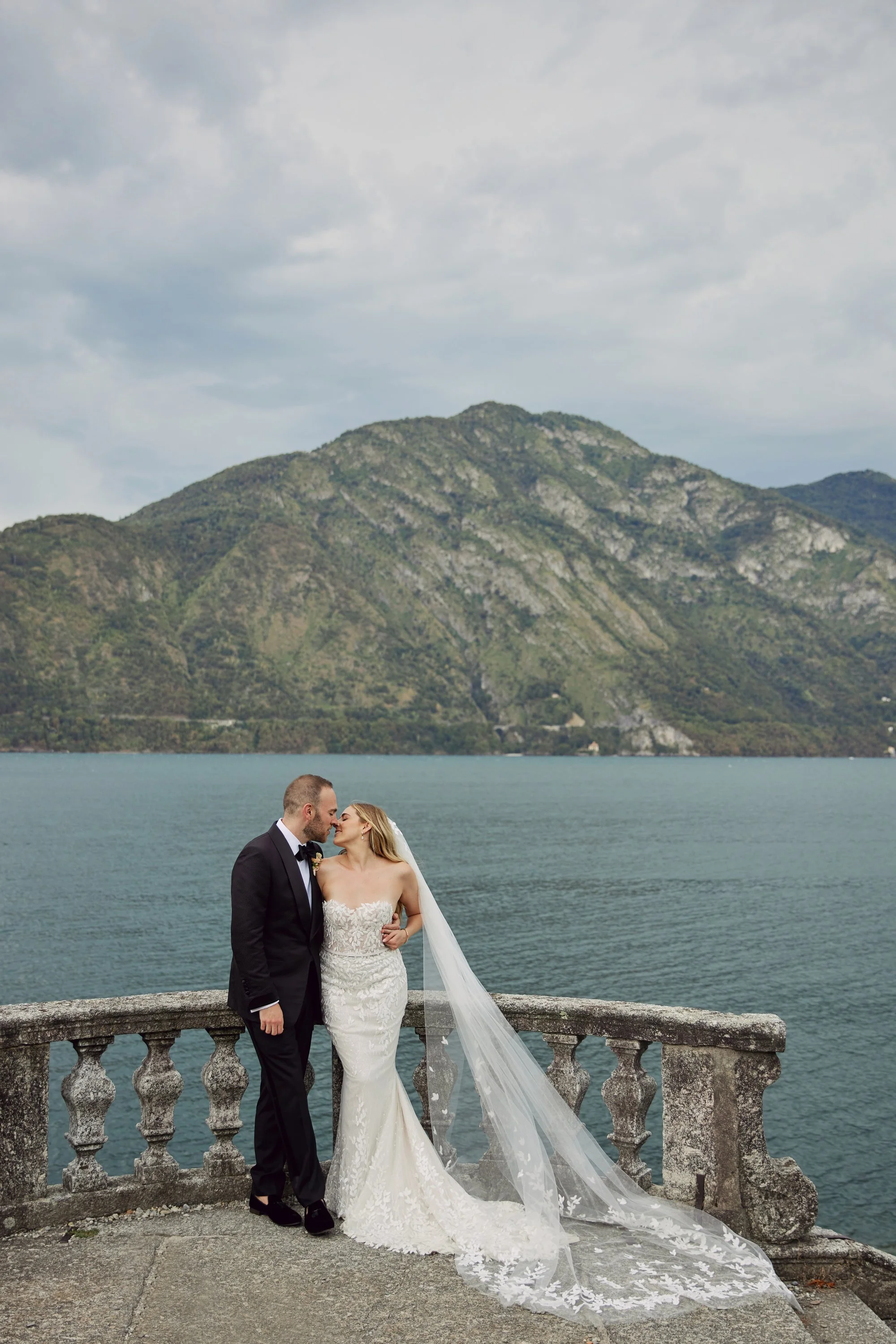 A newlywed couple stands close together on a stone balcony overlooking a lake with mountains in the background. The groom wears a black tuxedo, and the bride wears a white lace wedding gown with a long veil. They are touching foreheads and smiling.