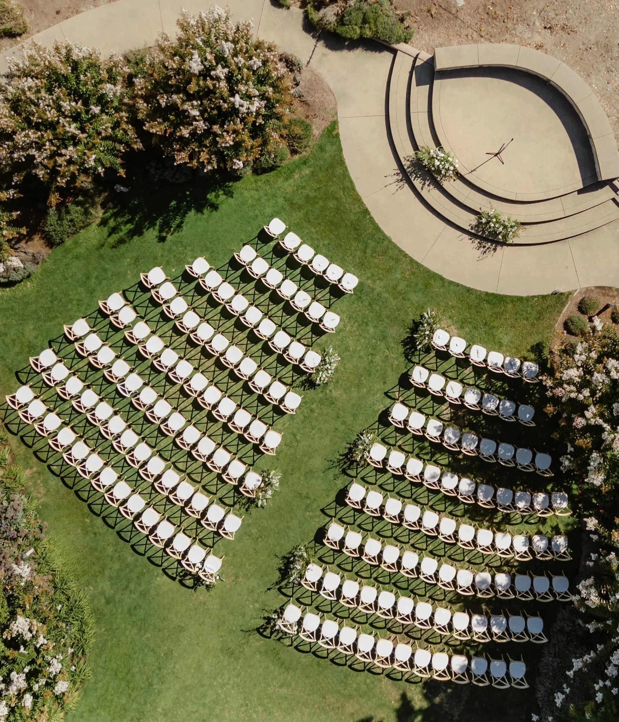 An outdoor wedding setup with rows of white chairs on a grassy lawn, a semi-circular concrete stage with floral arrangements, surrounded by blooming bushes and trees.