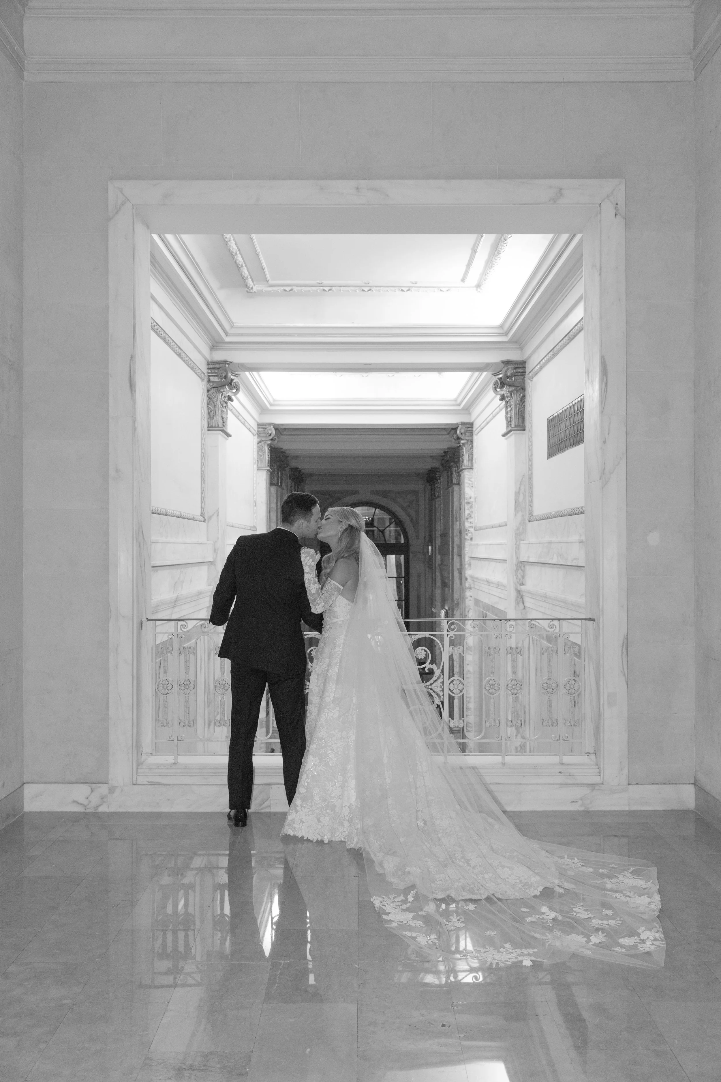 A newlywed couple sharing a kiss in a grand, marble-floored hallway with ornate architectural details, seen through a large doorway.