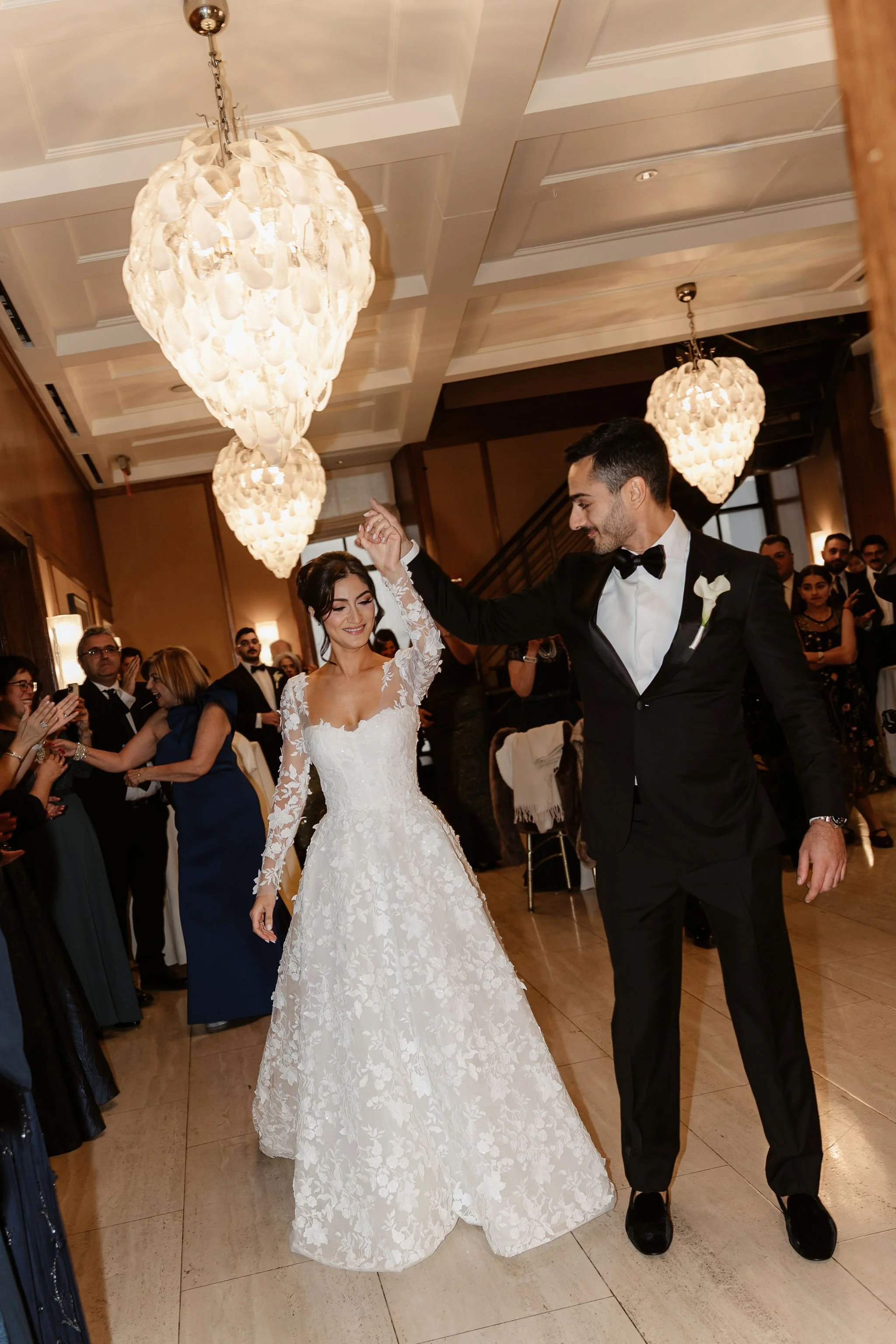 A bride and groom are dancing at their wedding reception, surrounded by guests. The bride is wearing a white lace wedding gown, and the groom is in a black tuxedo with a bow tie. The room is decorated with elegant chandeliers and warm lighting.