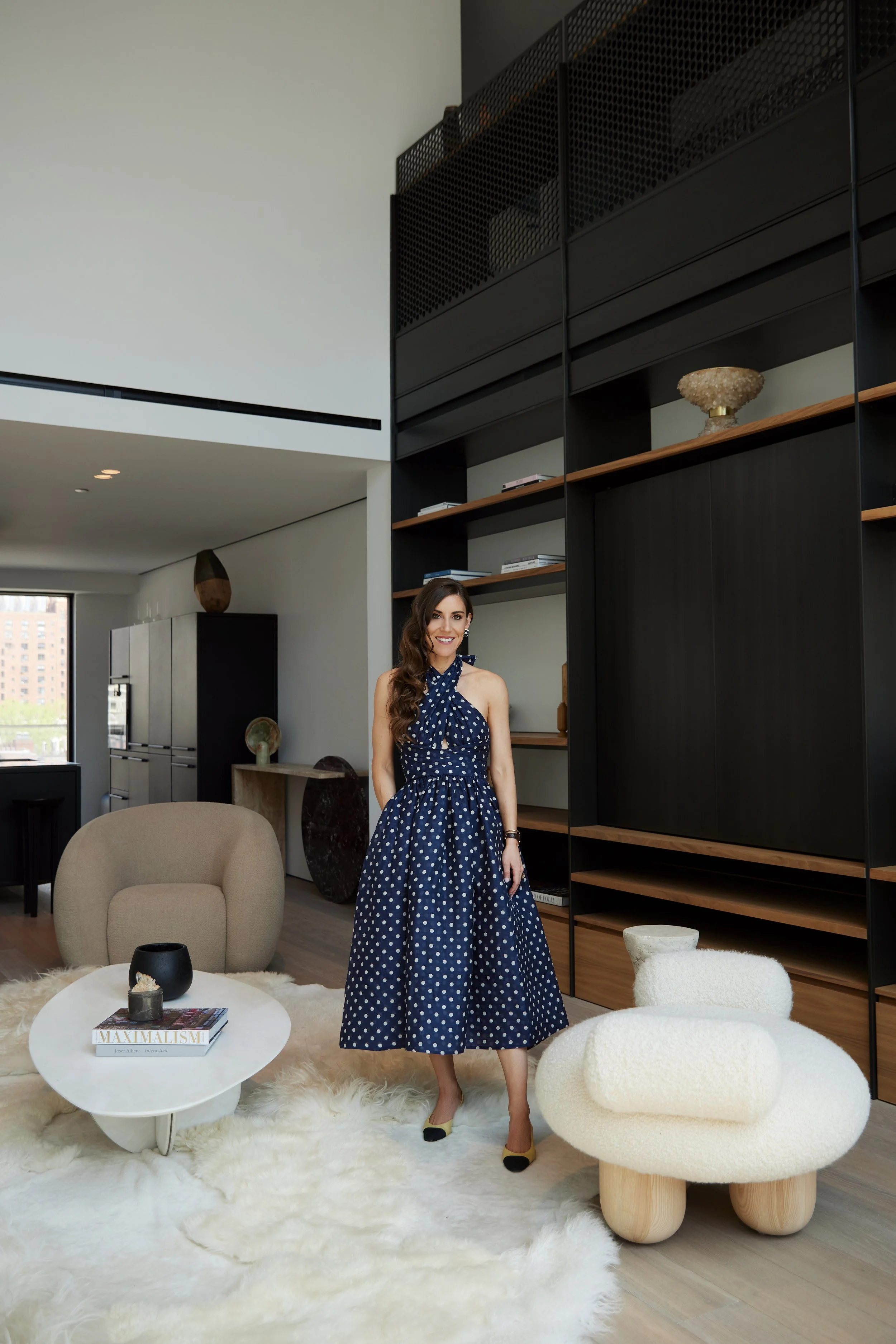 Woman in blue polka dot dress standing in a modern living room with white shaggy rug, beige armchair, white coffee table, and black shelving unit with decor items.