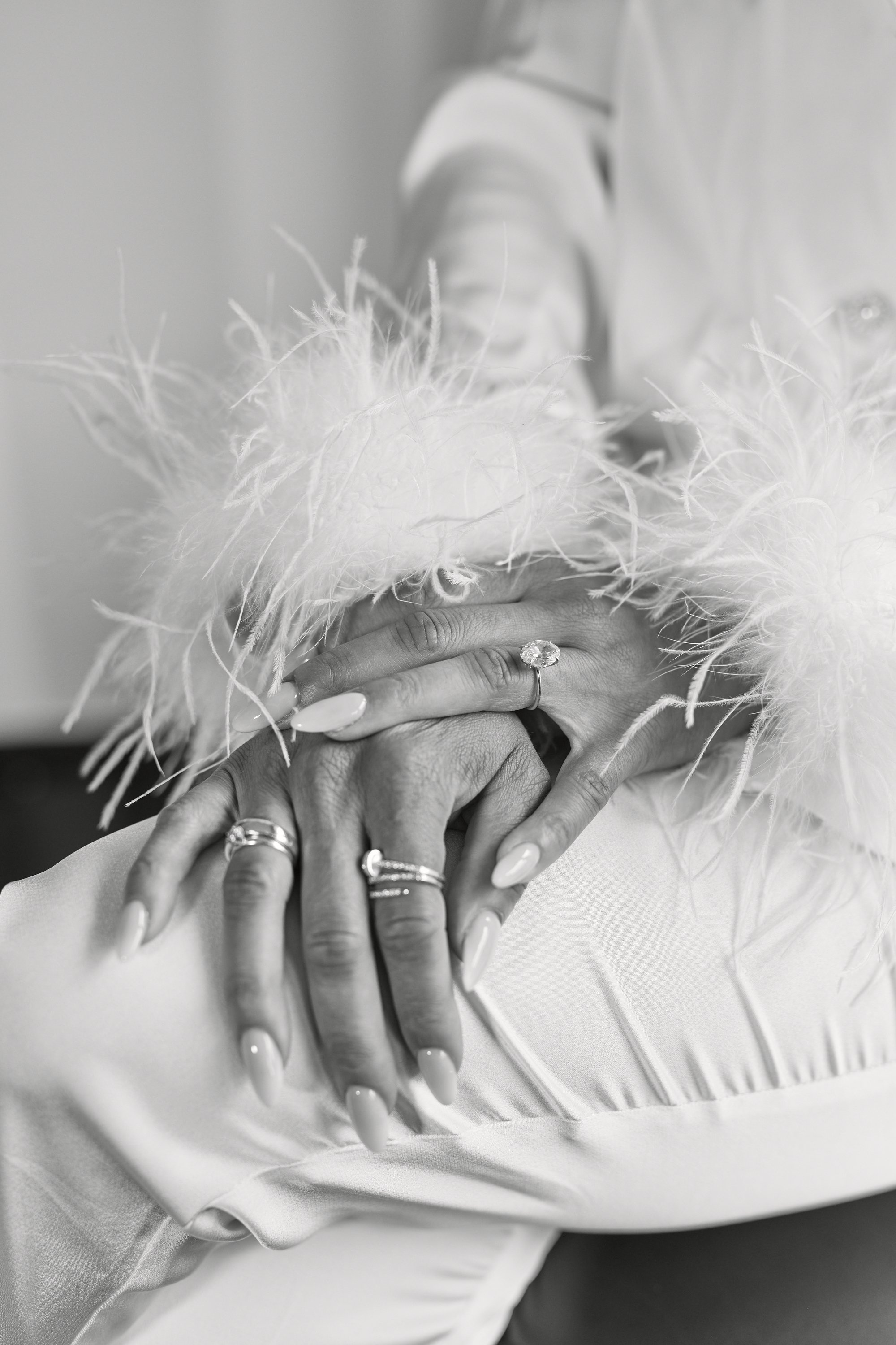 Close-up black-and-white photograph of a person's hands clasped together, wearing multiple rings and a feathered shawl, with a focus on the hands and rings.