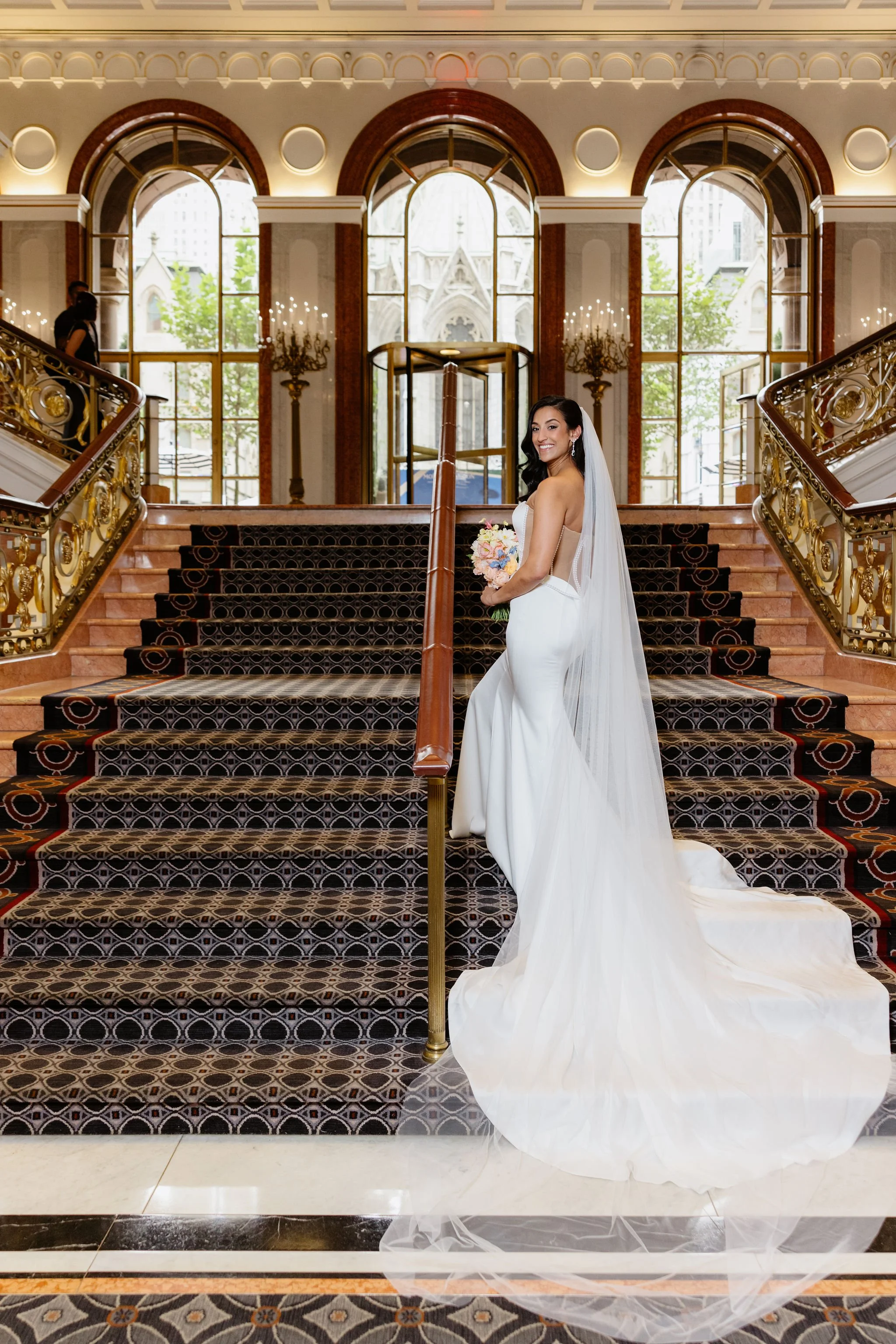 A bride in a white wedding dress holding a bouquet of pink and white flowers posing on a grand staircase with ornate gold railing in a luxurious building with large arched windows and decorative brass wall sconces.