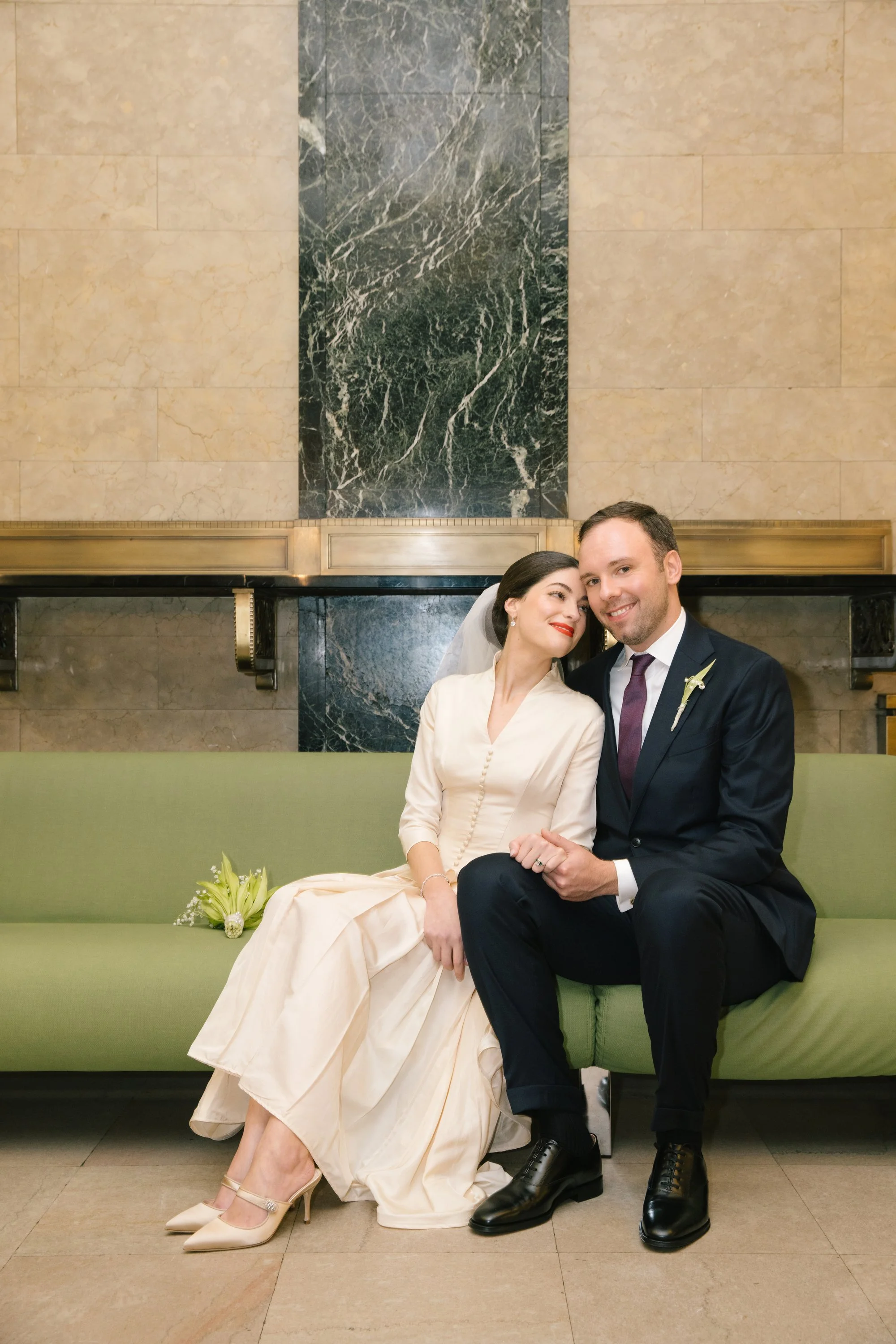 A newlywed couple sitting on a green sofa in front of a marble fireplace. The bride is in a light-colored dress, and the groom is in a dark suit. They are smiling and leaning towards each other, with the bride resting her head on the groom's shoulder. A bouquet of flowers is placed on the sofa beside the bride.