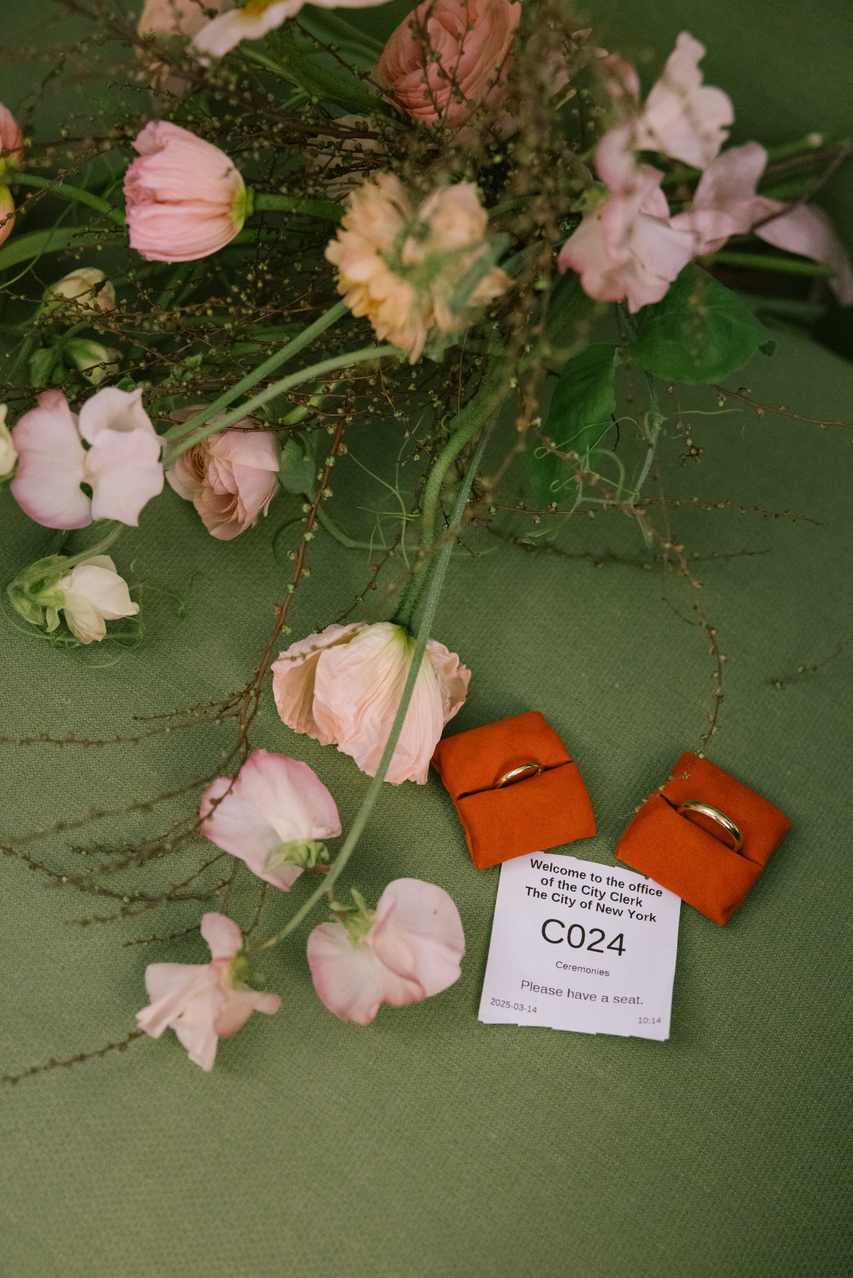 A floral arrangement of pink and white flowers with green leaves on a green surface, accompanied by orange rings and a note indicating it is for a ceremony at the City of New York.