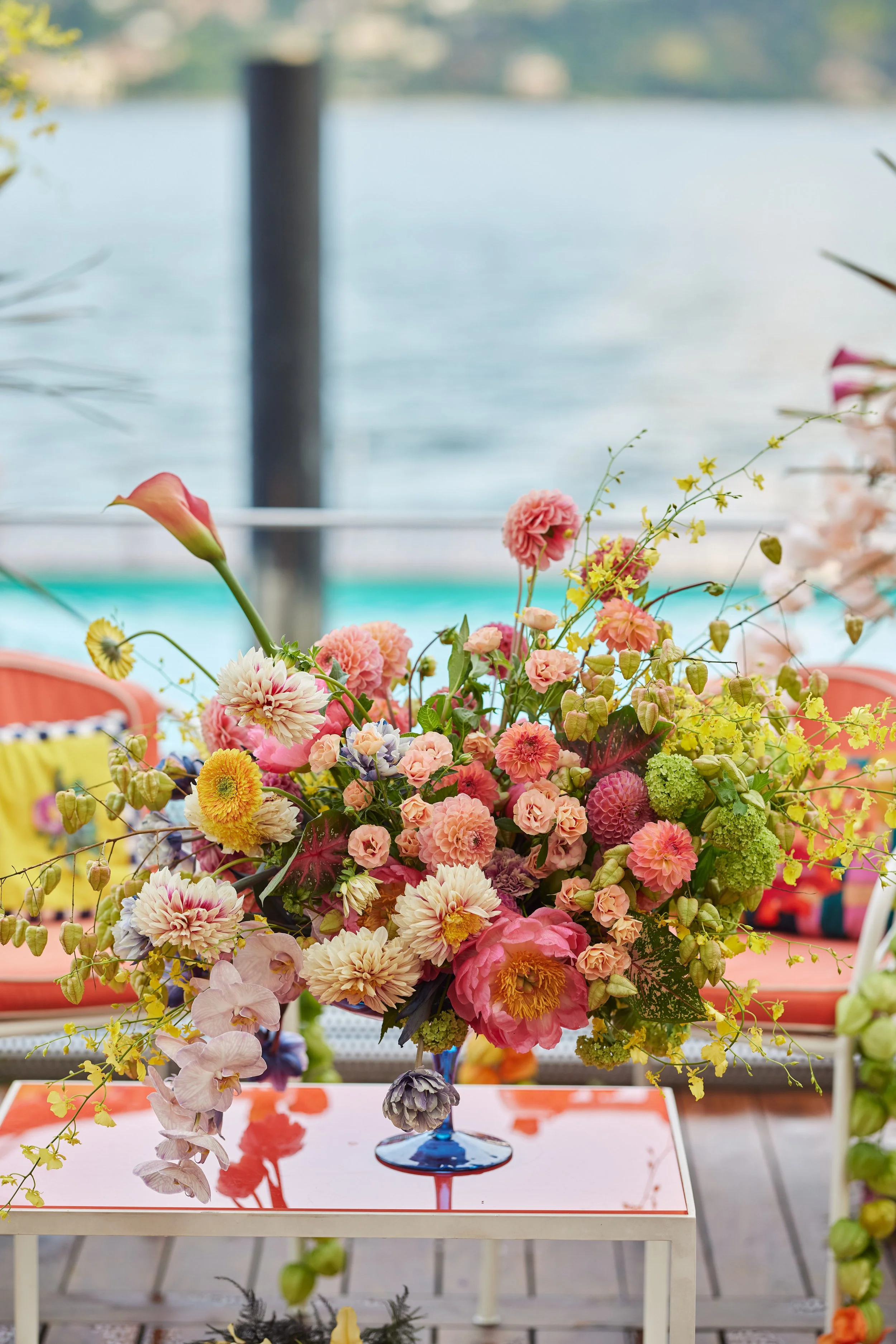 A vibrant floral arrangement in a blue glass vase placed on a white table with a mirror surface, set outdoors near a body of water with a blurred background of trees and water.