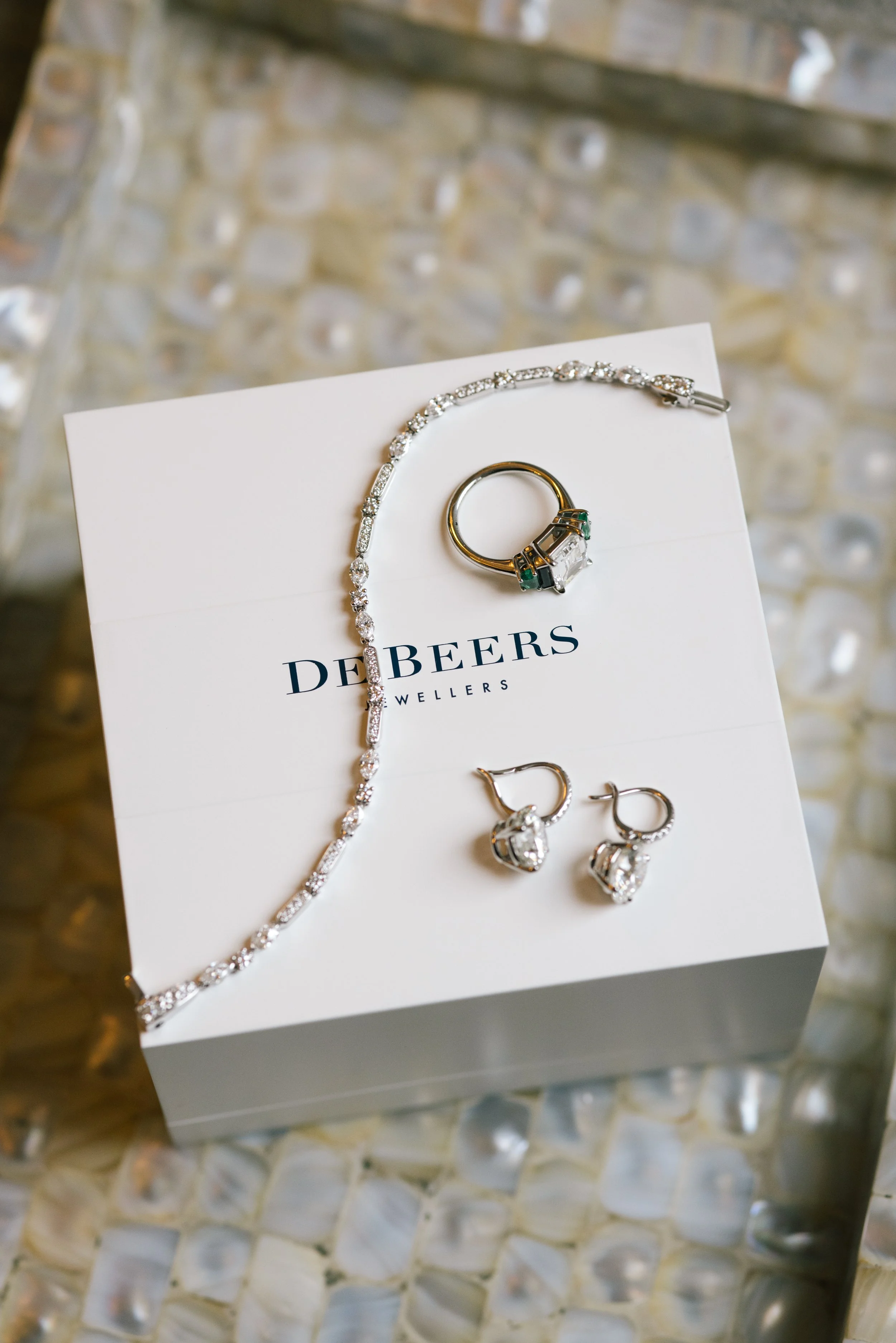 Jewelry display on a white card with the brand name 'De Beers'. The jewelry includes a diamond necklace, a ring with green accents and clear stones, and matching earrings, set on a textured, glittery background.