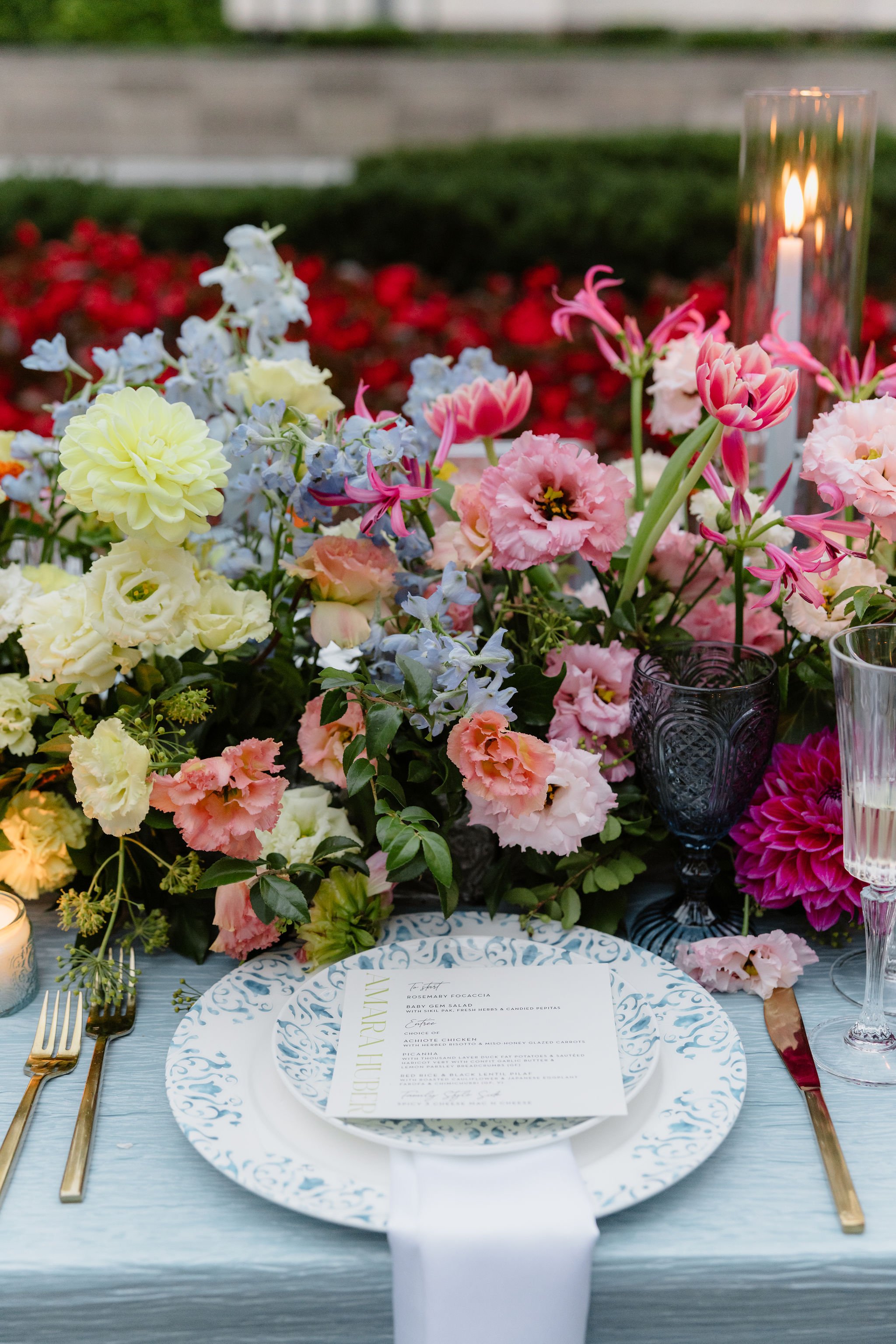 A table setting with a floral centerpiece of pink, white, and yellow flowers, a white plate with a detailed blue rim, a party menu, gold utensils, a dark glass goblet, and a lit candle in a tall glass holder.