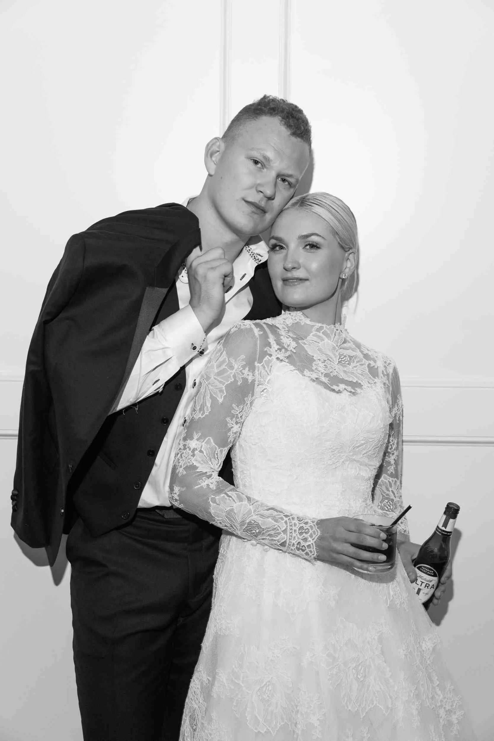 A young man and woman dressed in wedding attire posing together in front of a plain background, with the woman holding a drink and a bottle of beer on the table.