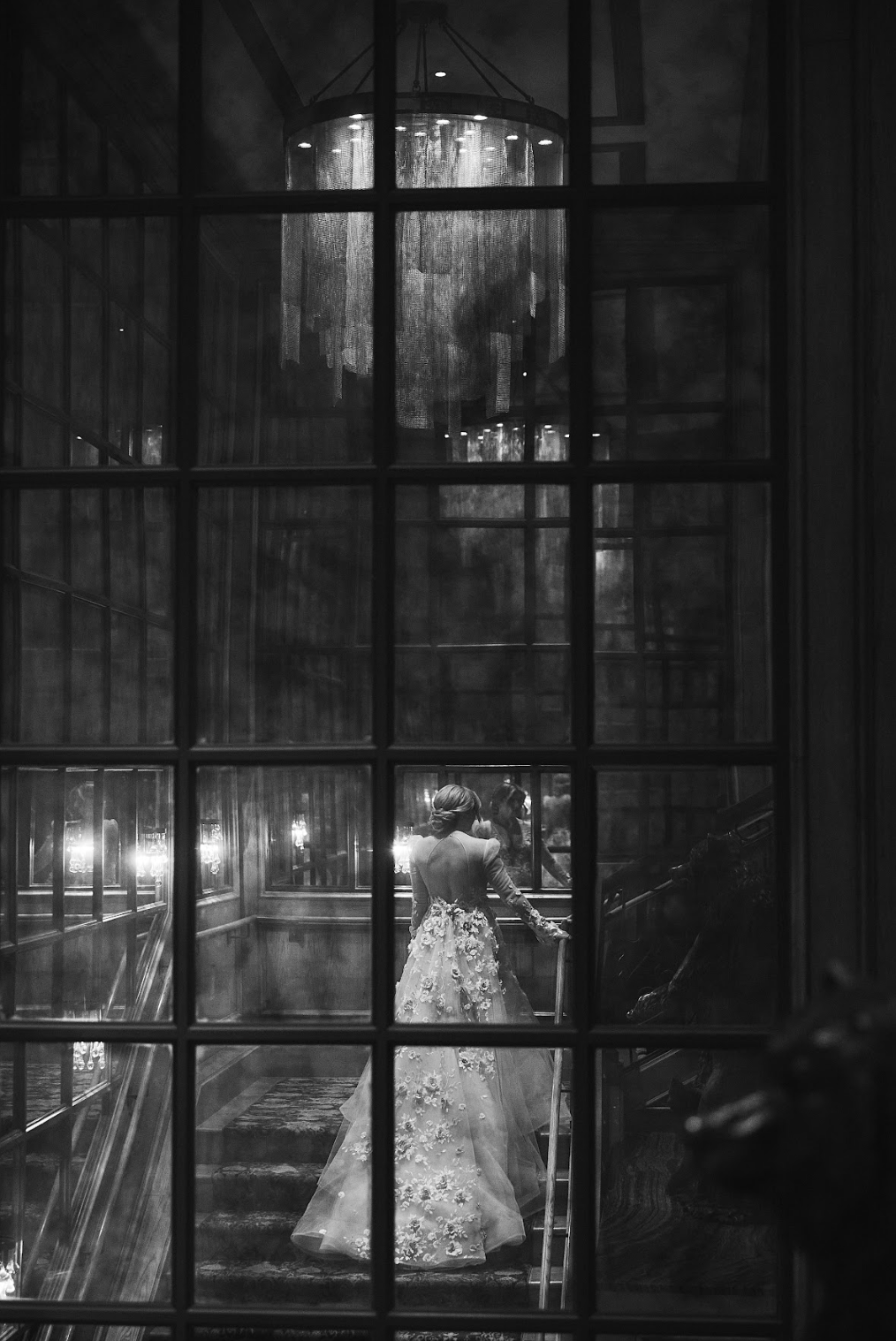 Black and white photo of a woman in a wedding dress walking up stairs, viewed through a window with grid panes.