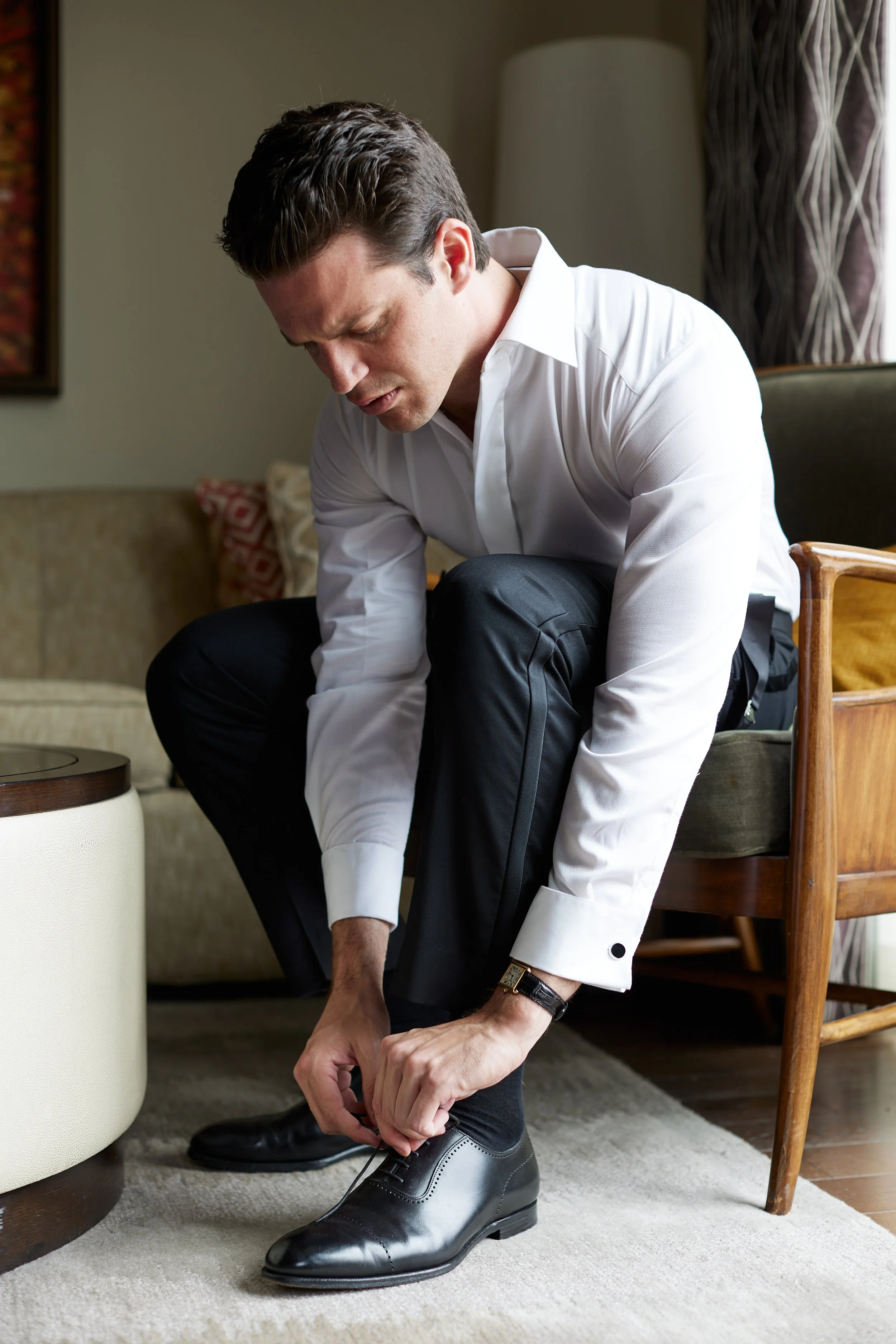 A man dressed in formal attire, sitting on a chair, tying his shoelace, in a living room setting.